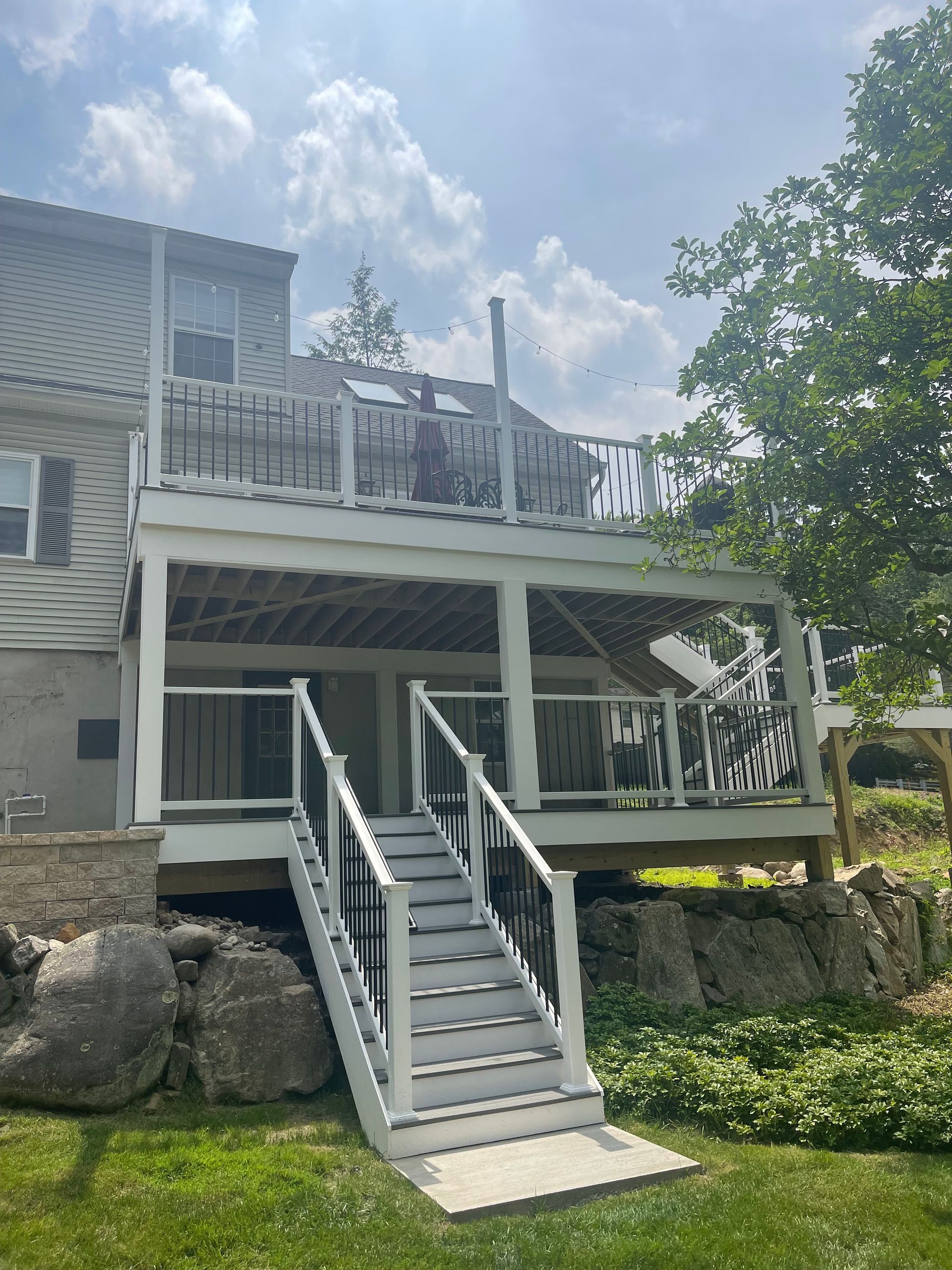Two-story white deck with black railing against a house with stone accents, stairs leading to the yard on a sunny day.