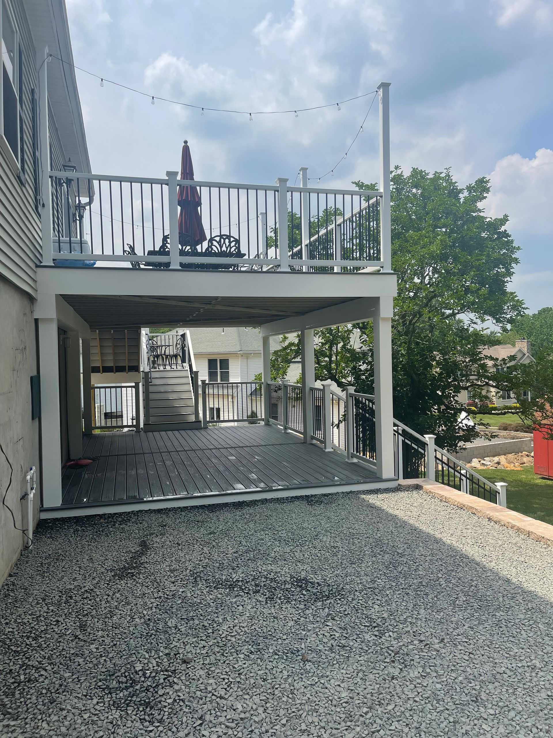 Two-story deck with gravel below; upper deck with railing, umbrella, and lights; house to the left.