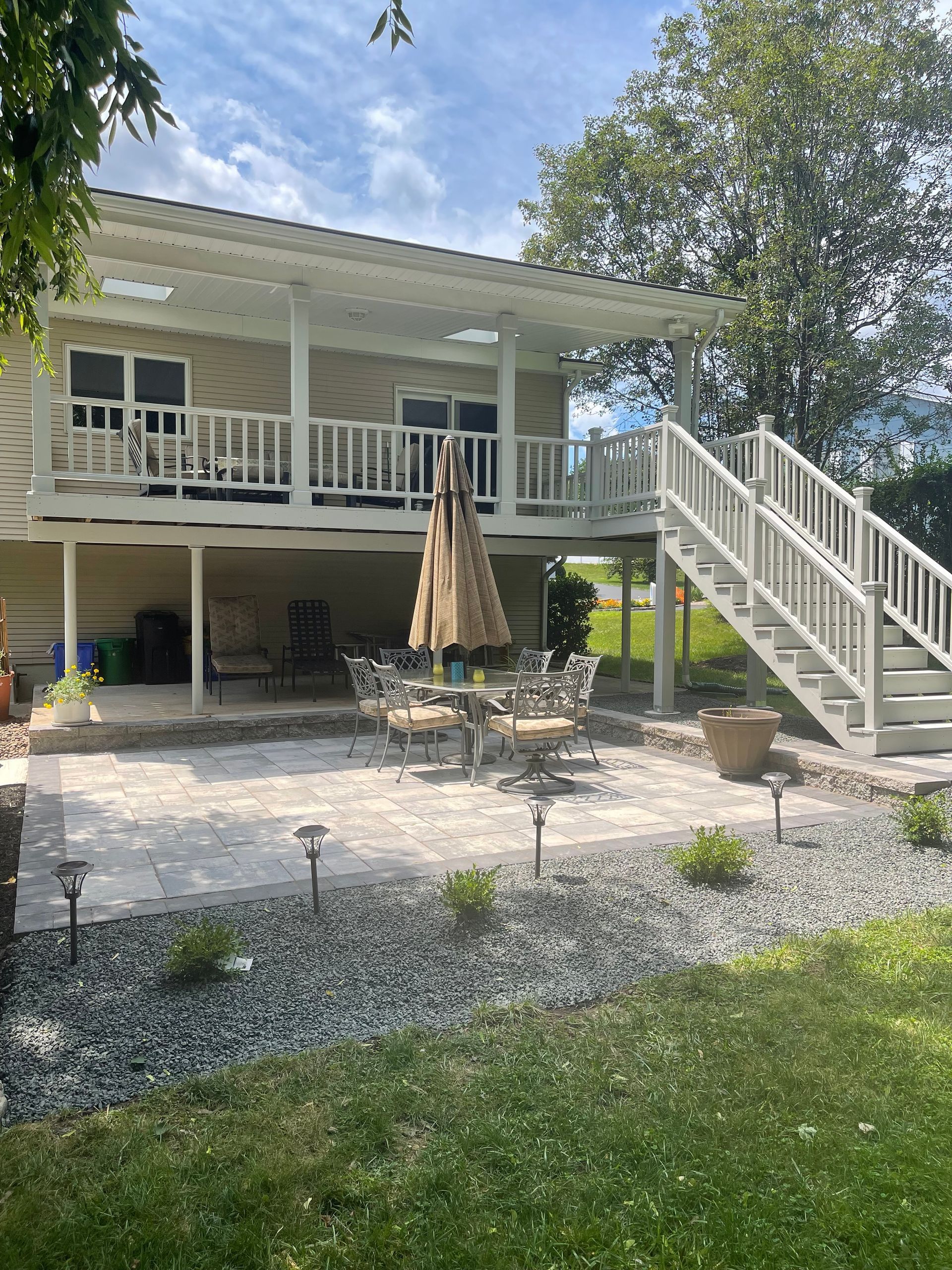 Patio with stone pavers, table, chairs, and raised deck with stairs, next to a house under a sunny sky.