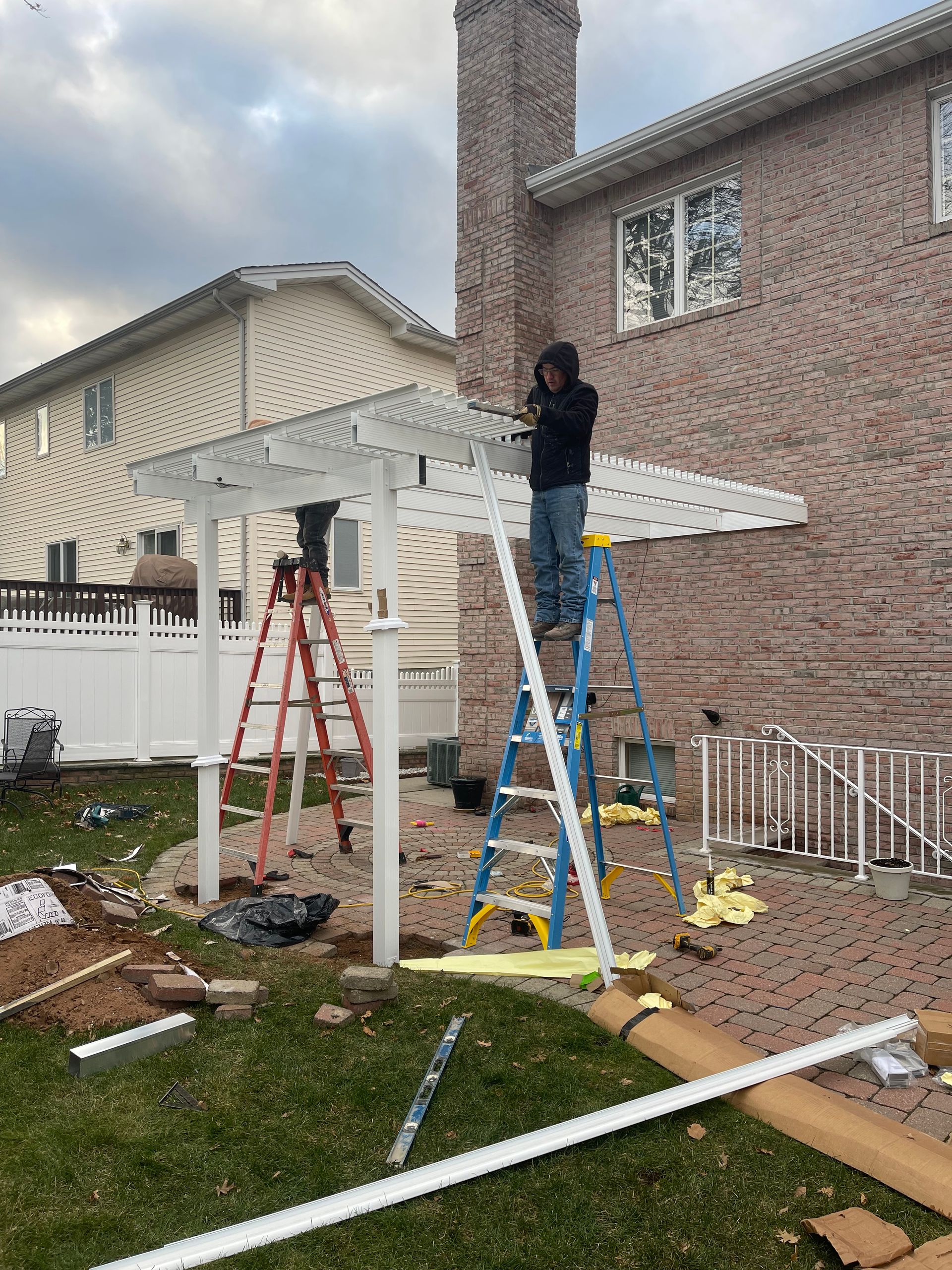 Two workers installing a white pergola next to a brick house. One stands on a blue ladder.
