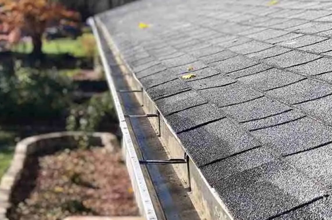 Gutters along a gray shingled roof, filled with leaves.