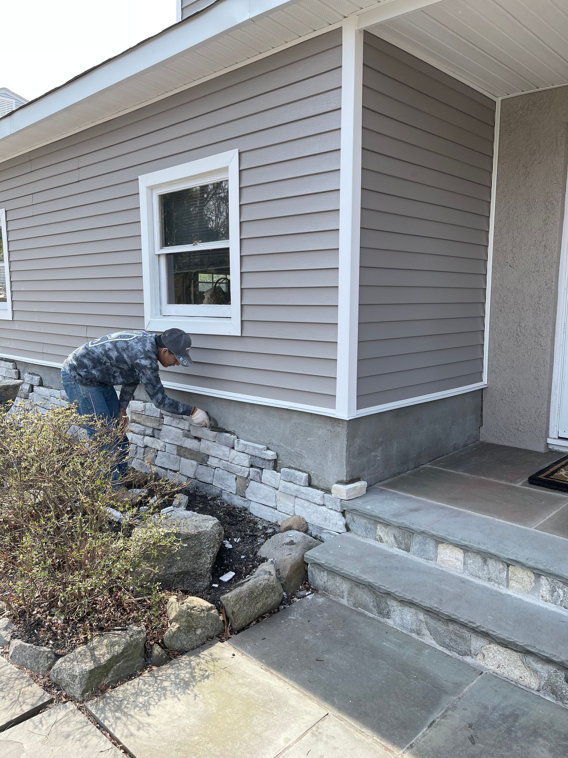 Person working on exterior brick siding of a house with gray siding and white trim.