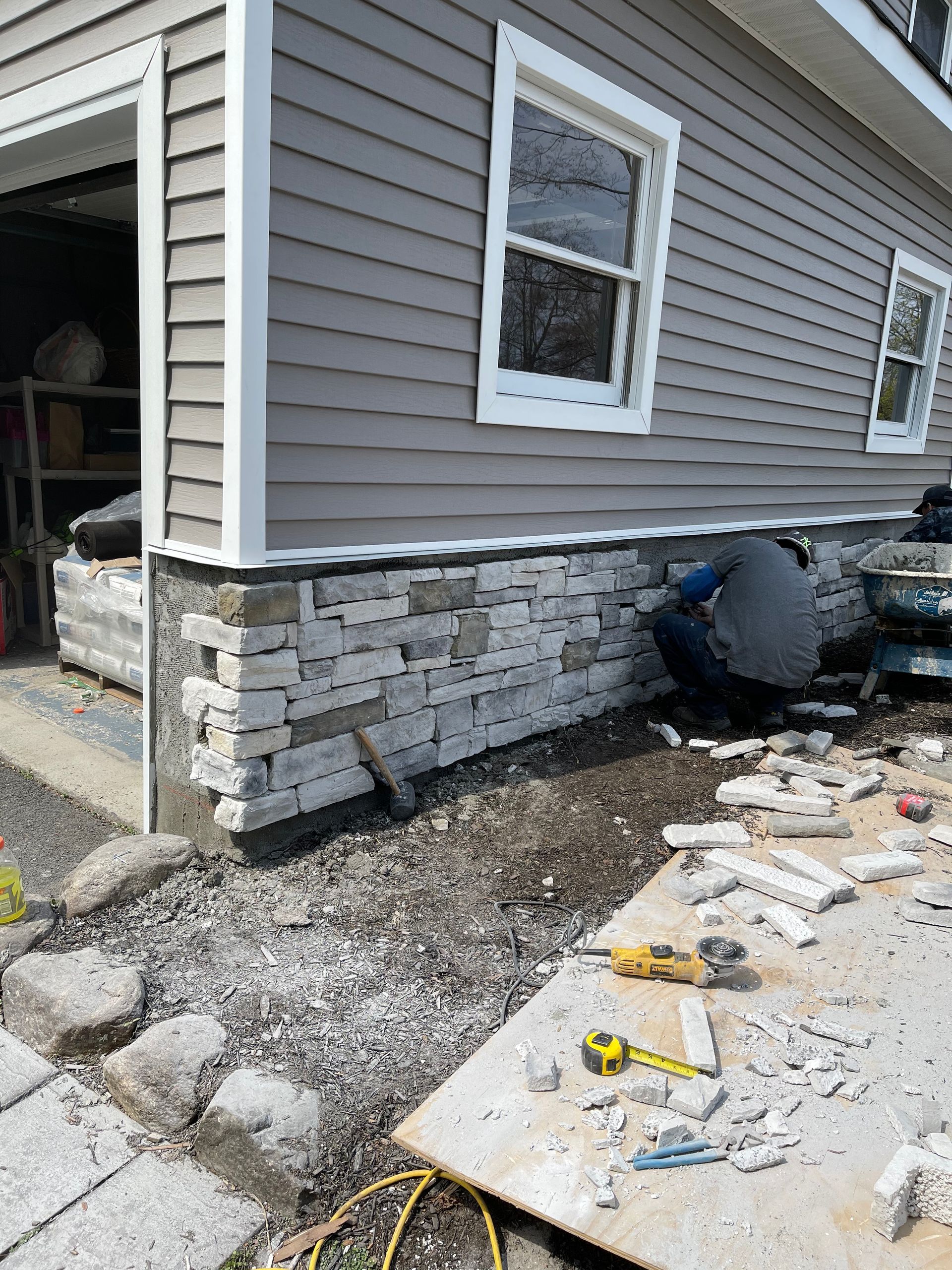 Stone veneer installation on a building's foundation. Person working on the project; siding and windows visible.