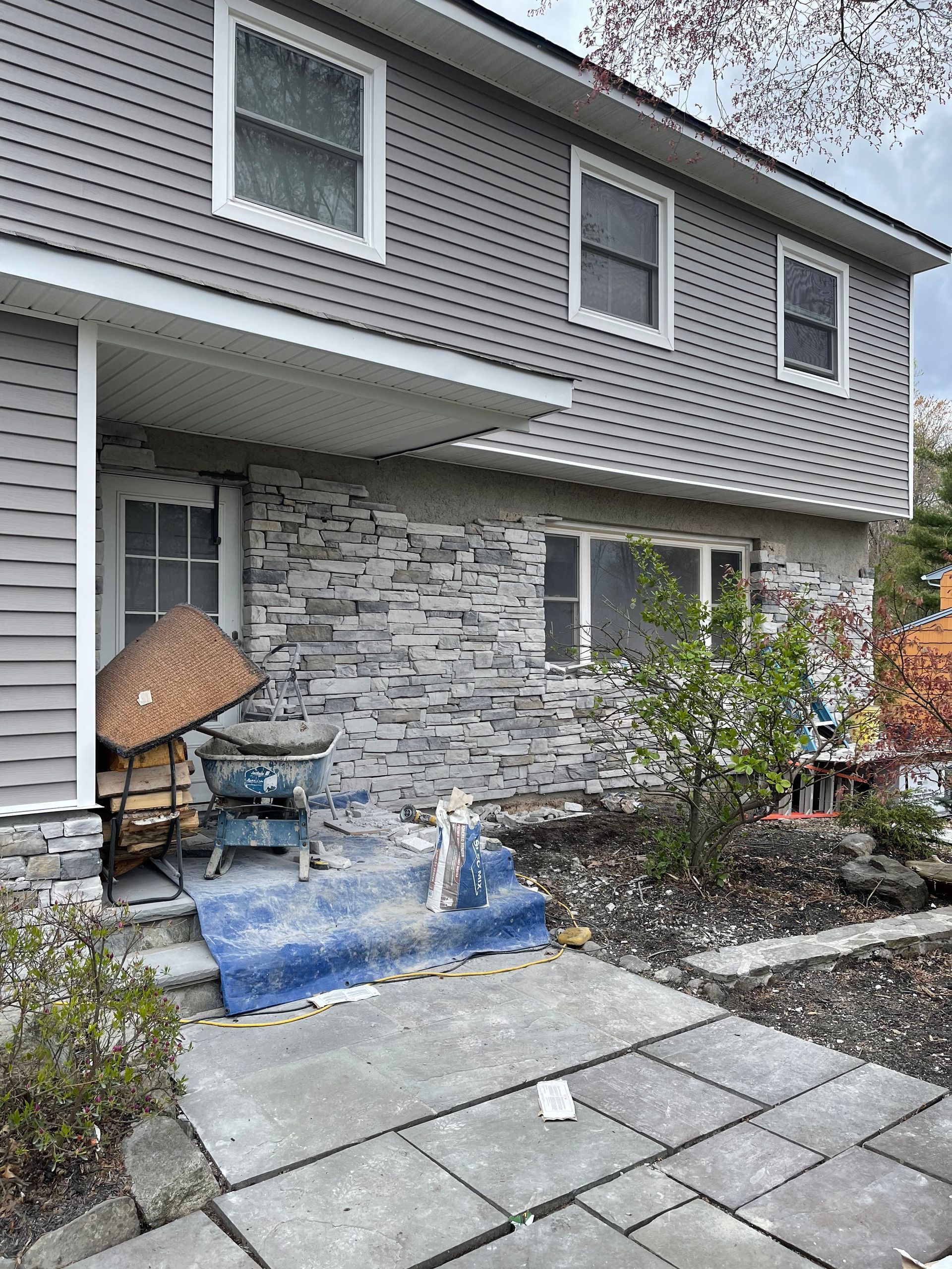 Two-story house with gray siding, stone facade on the porch, and a blue tarp on the front steps.