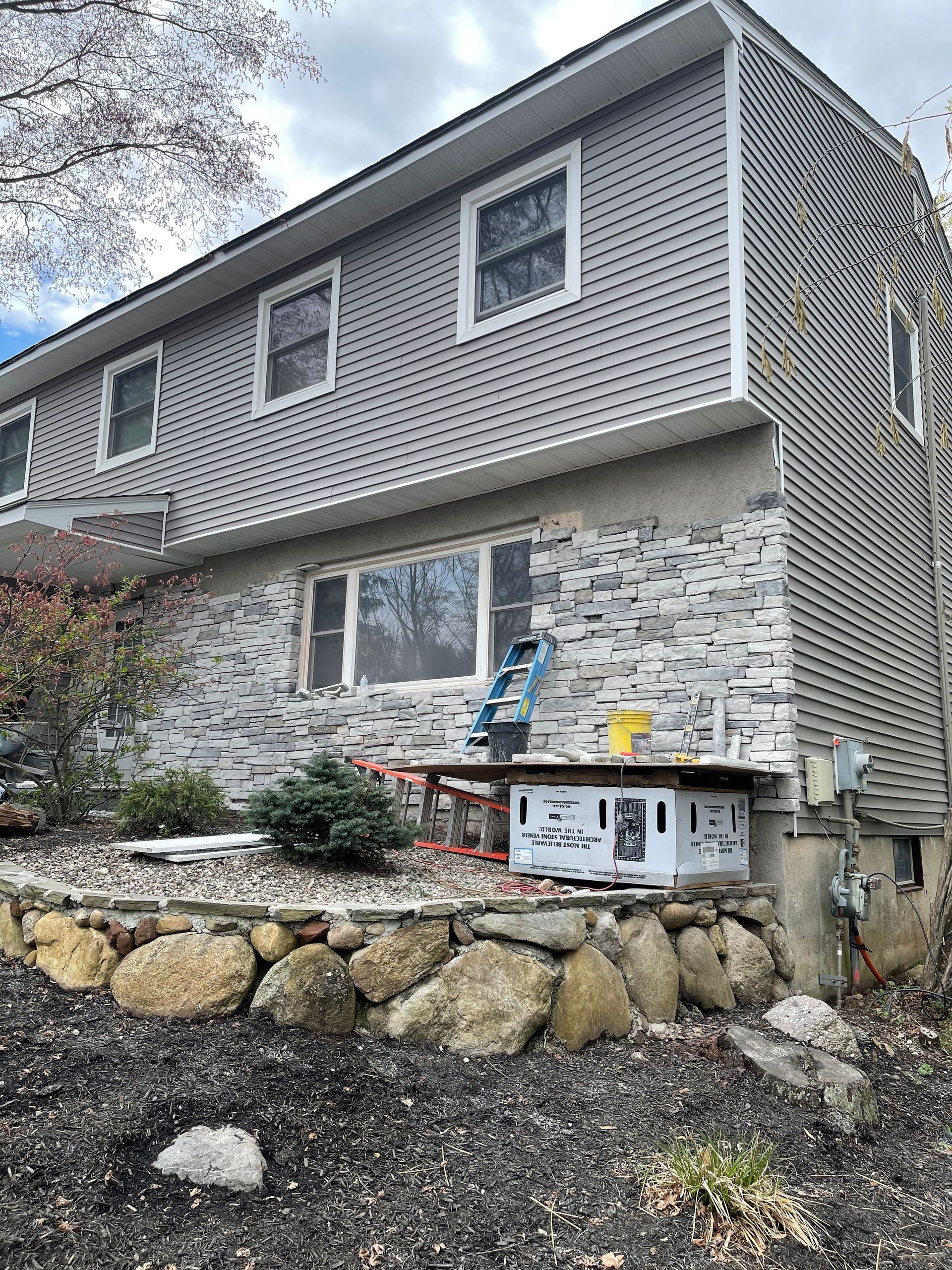 House with gray siding and stone facade. A ladder and tools are set up for exterior work.
