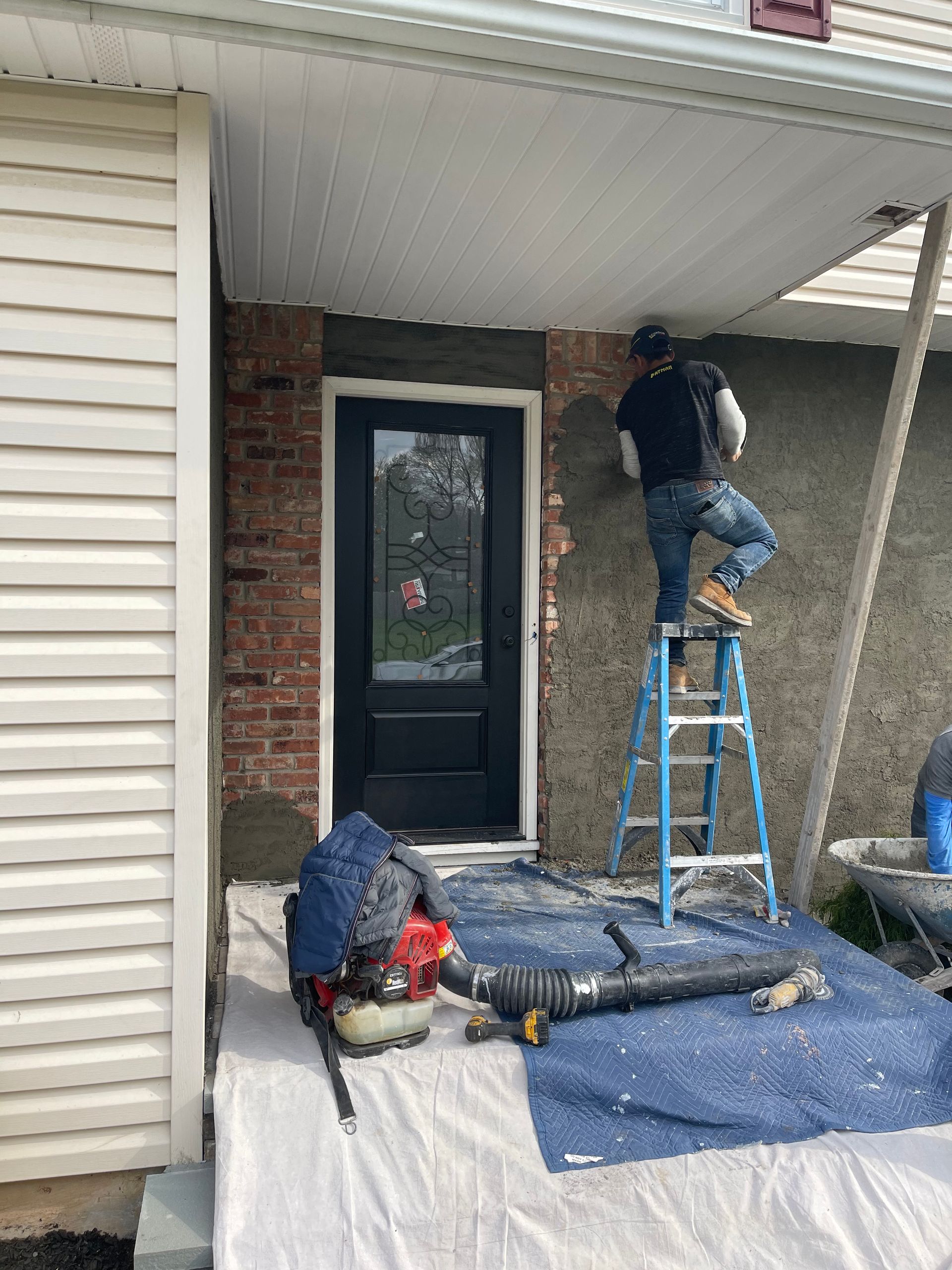 A construction worker applies stucco to a wall above a doorway, standing on a ladder. A leaf blower and supplies are nearby.
