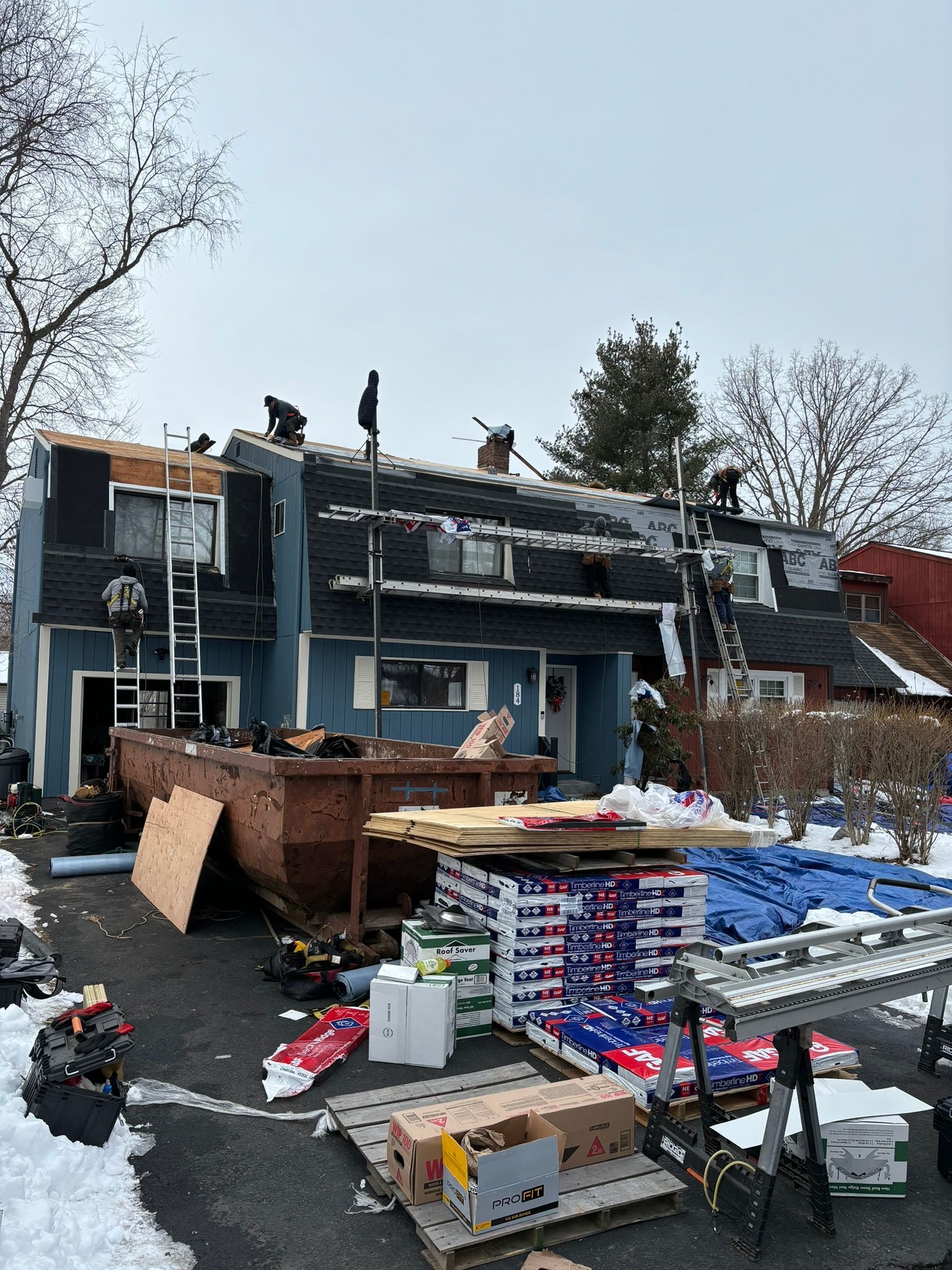 Roofers working on a blue multi-unit building in winter; ladders, dumpster, and supplies are present.