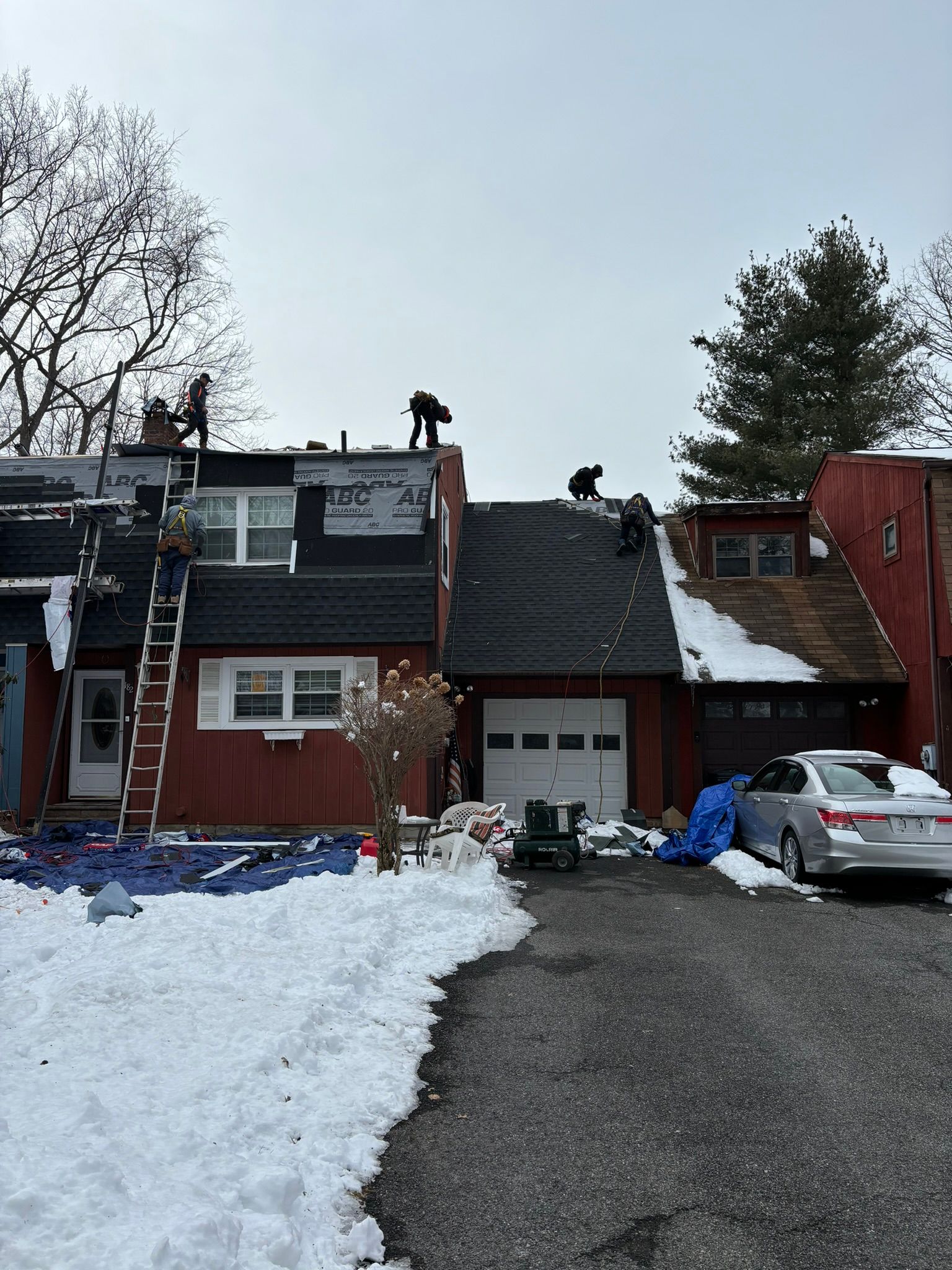 Roofers replacing shingles on a red townhouse. Snow on ground, ladder propped on the side. Overcast sky.