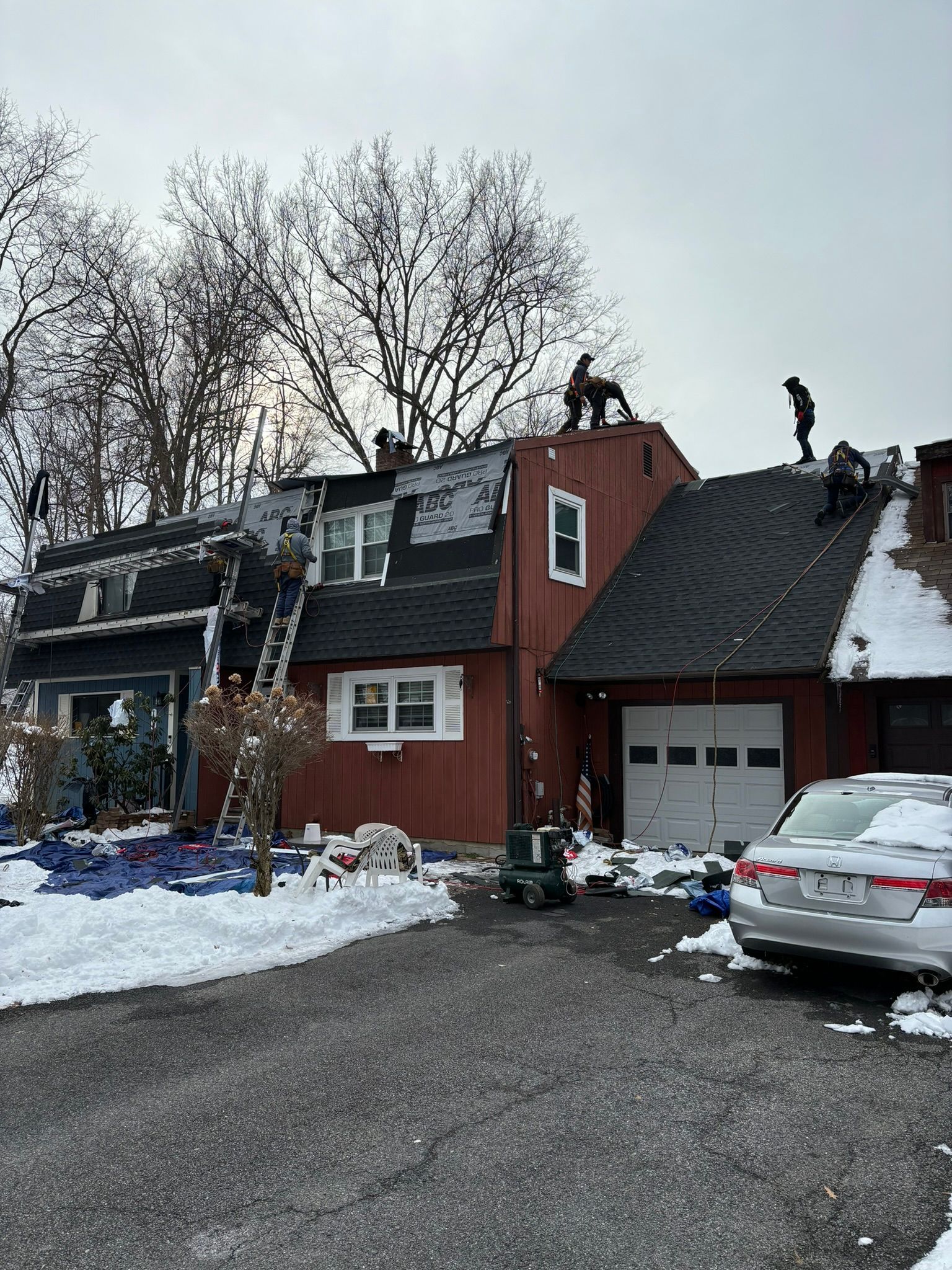 Workers on a roof during winter. Red house with snow, ladders, and a white garage door.