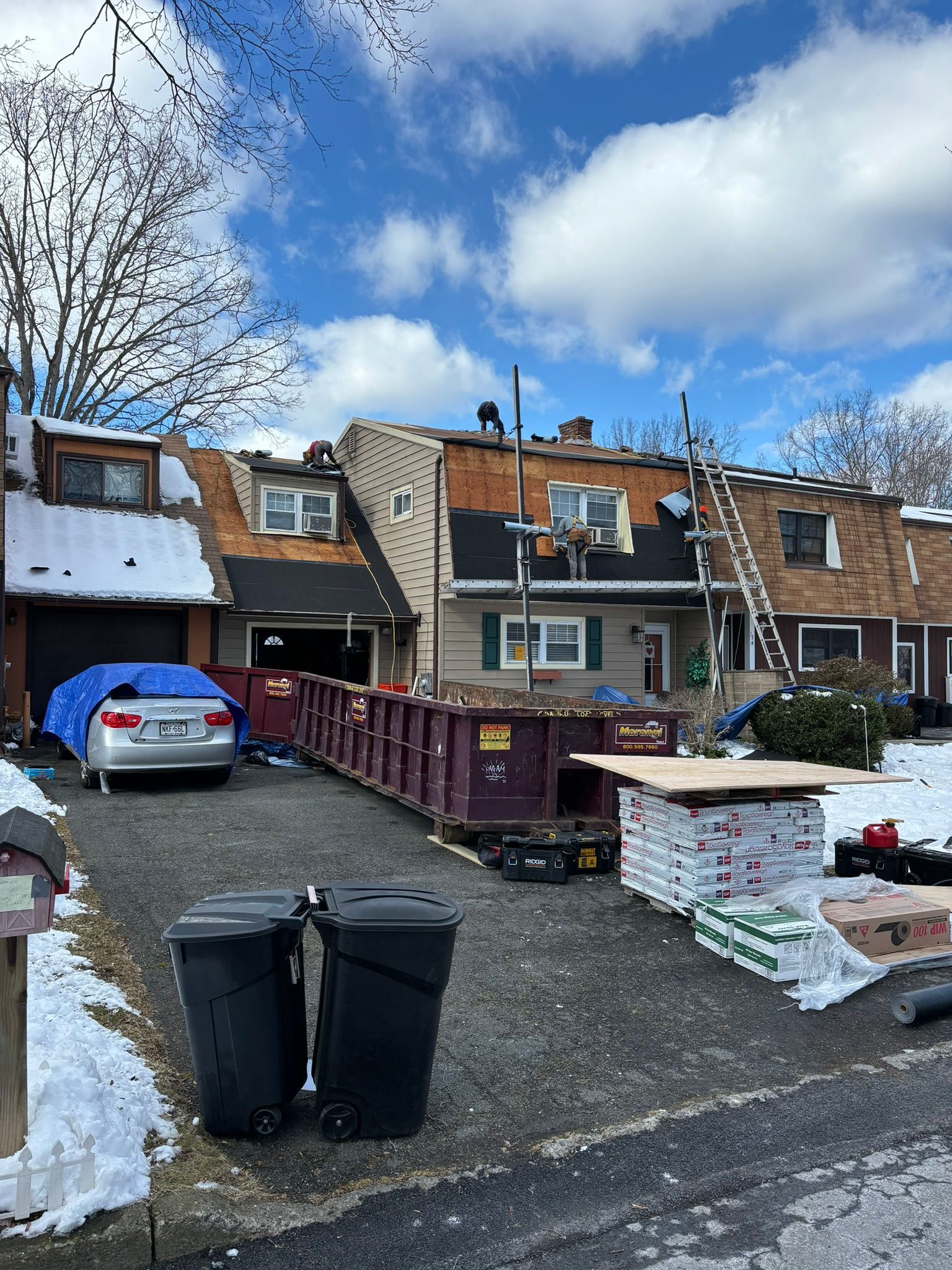 Residential building with roof replacement in progress. Dumpster, materials, and ladder present on driveway with snow.