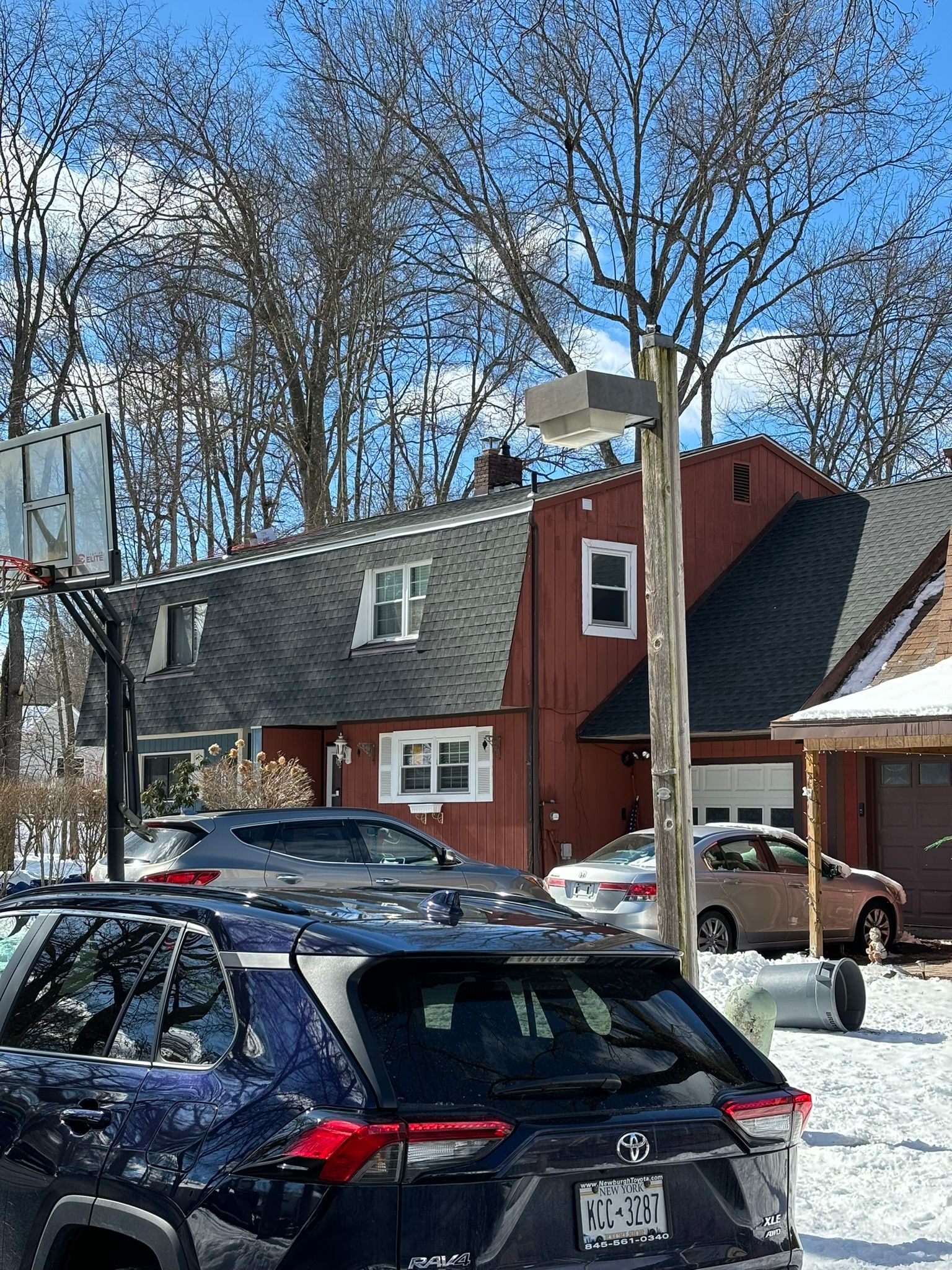 Red house with dark roof and parked cars in snowy yard. Basketball hoop. Street light.