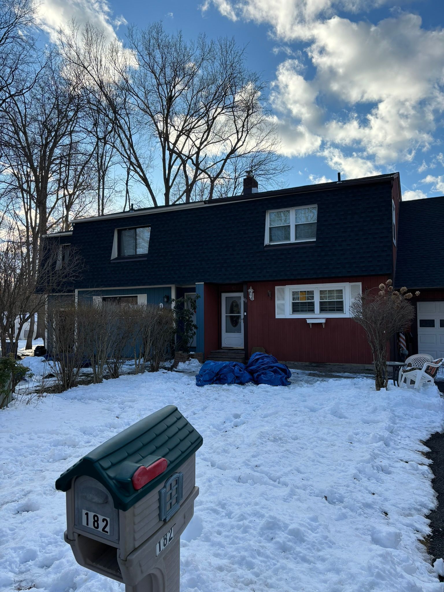 House with snow-covered lawn, mailbox in the foreground, and blue tarps near the entrance.
