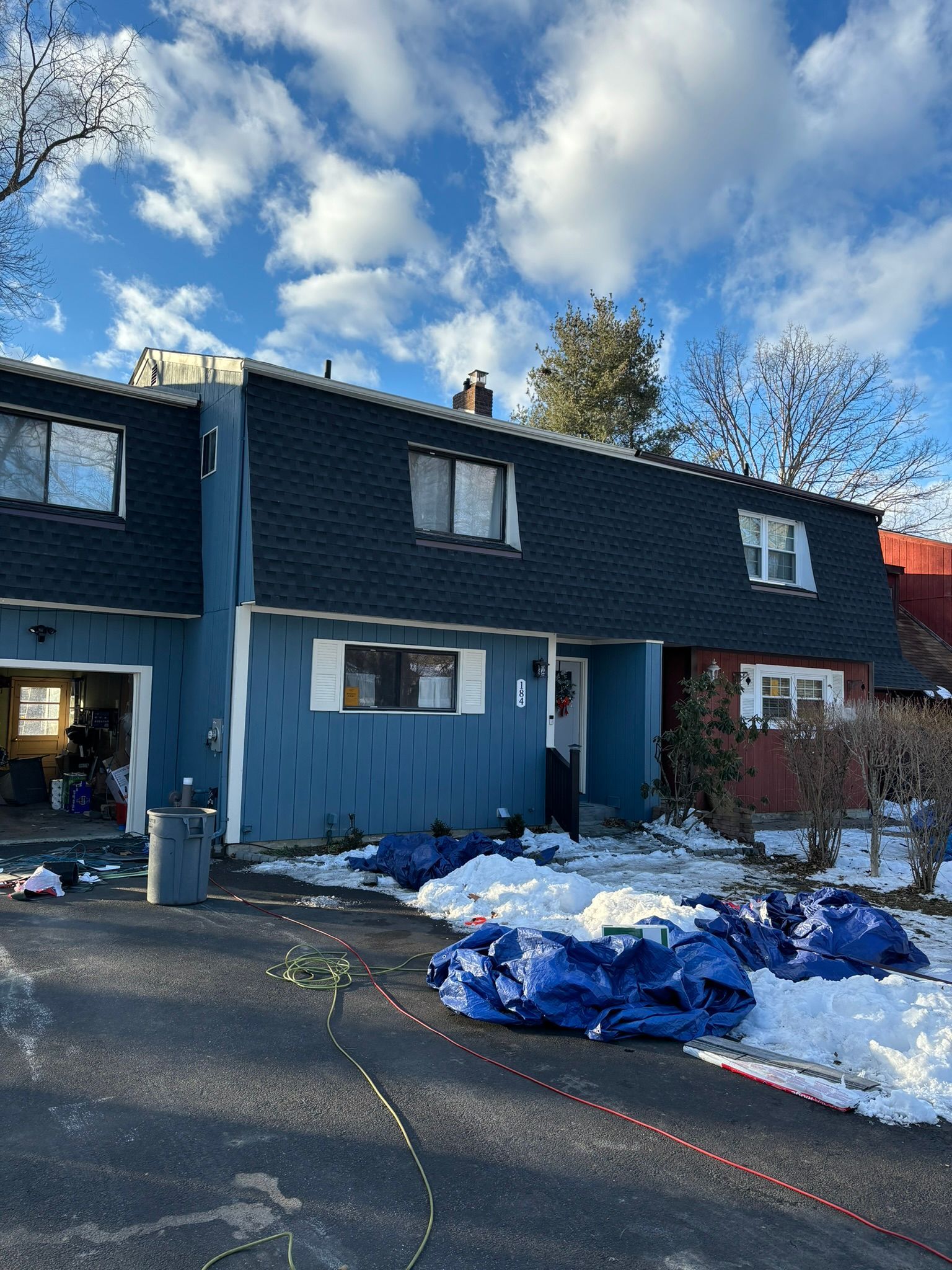 Townhouses with blue siding and dark roof, snow and blue tarps in front under a partly cloudy sky.
