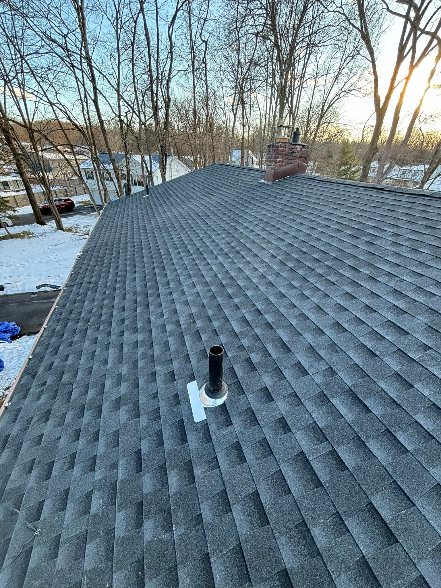 Overhead view of a gray shingled roof with a chimney and vent pipe, surrounded by bare trees and snow.