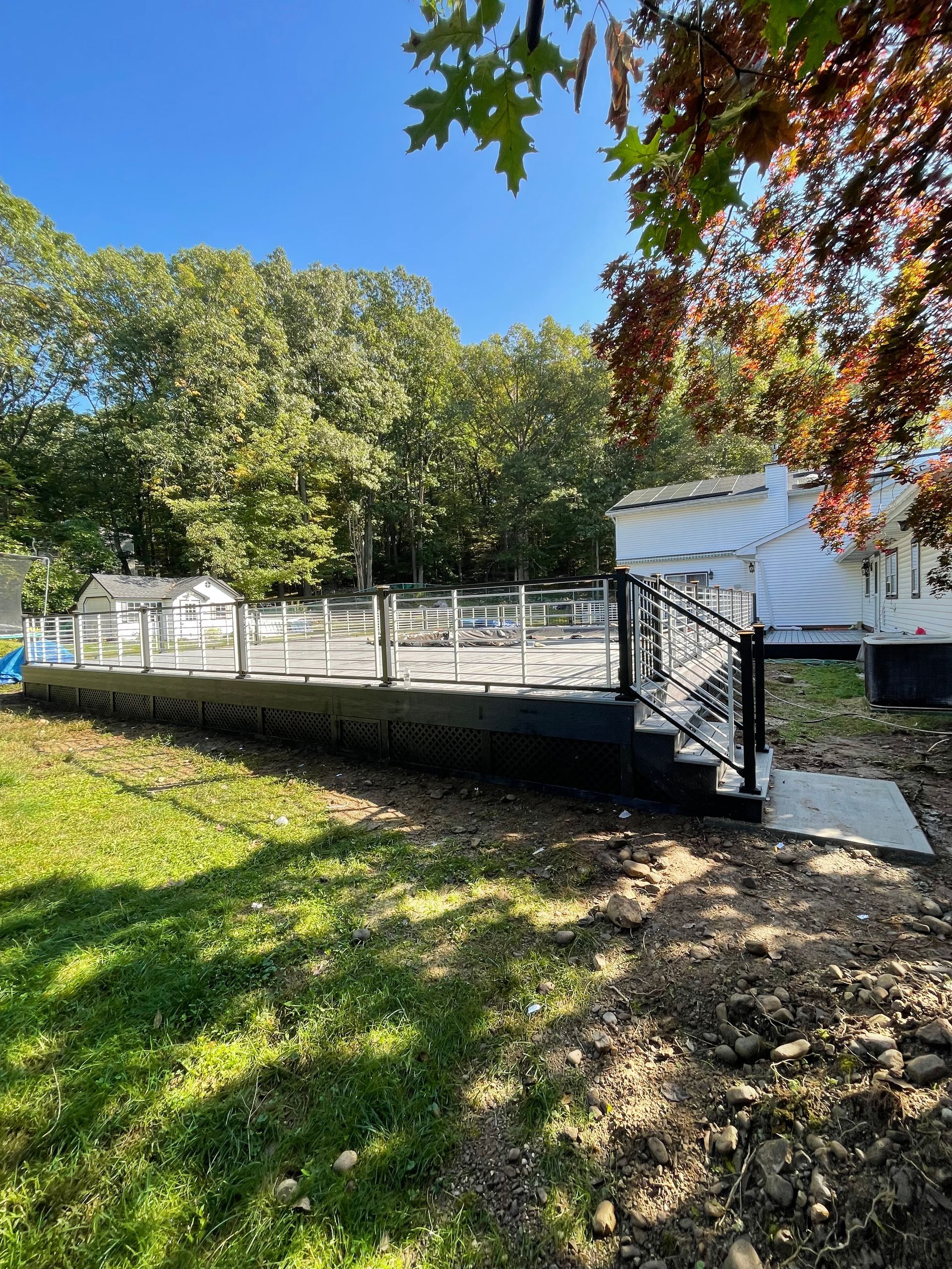 Deck with clear glass railing next to a white house, surrounded by trees and grass, on a sunny day.