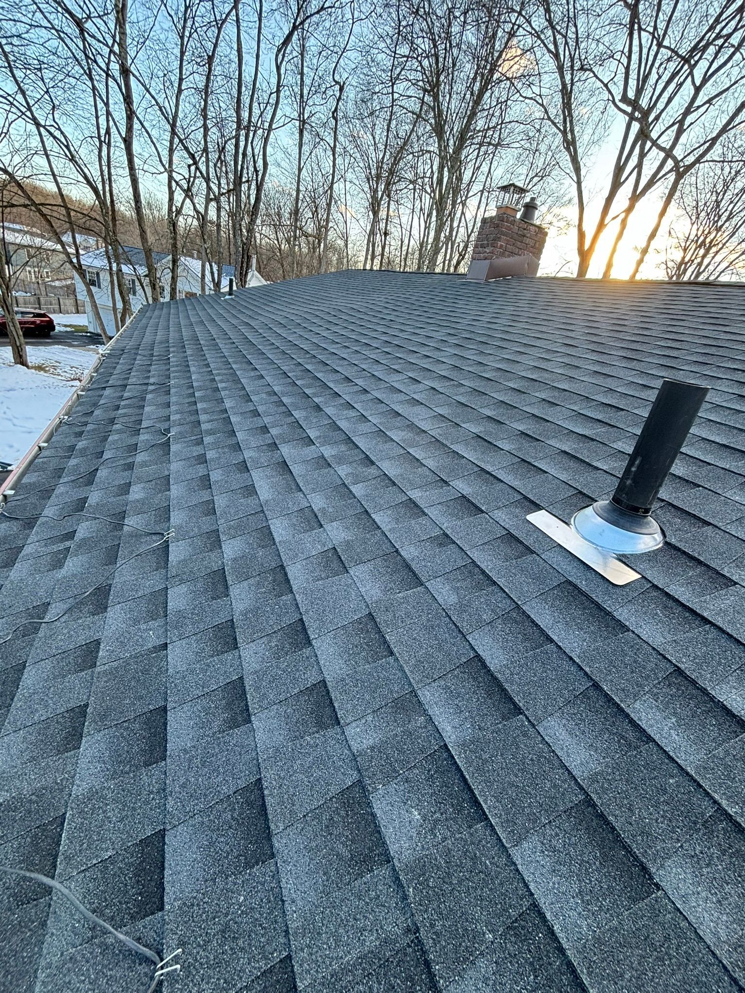 Asphalt shingle roof with a vent pipe and chimney, snow on the shingles. Bare trees and sky in the background.
