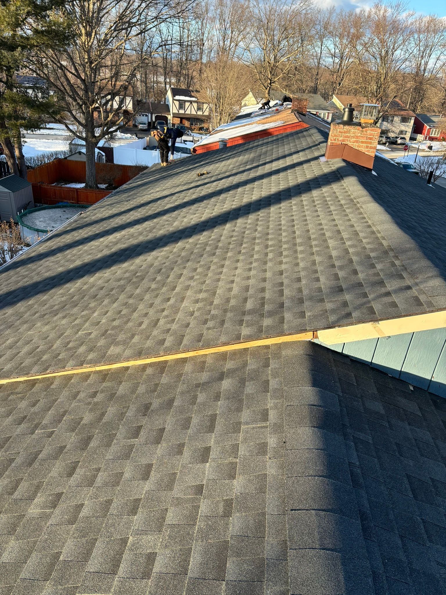 Roof being shingled; workers on the rooftop. Gray shingles, orange and brown trim, sunny day.