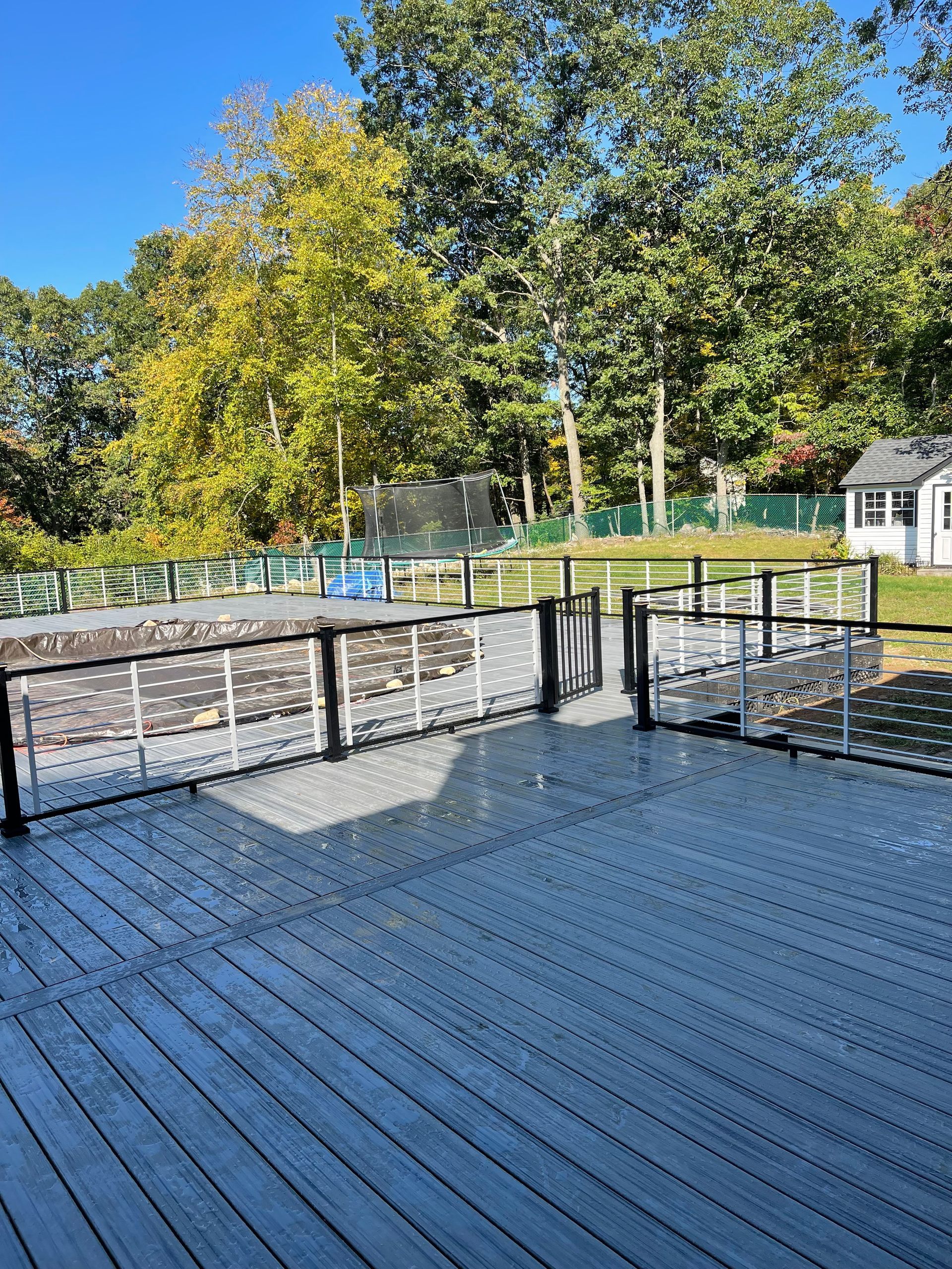 Gray deck with fence, pool in the background, trees, and a small white building under a blue sky.