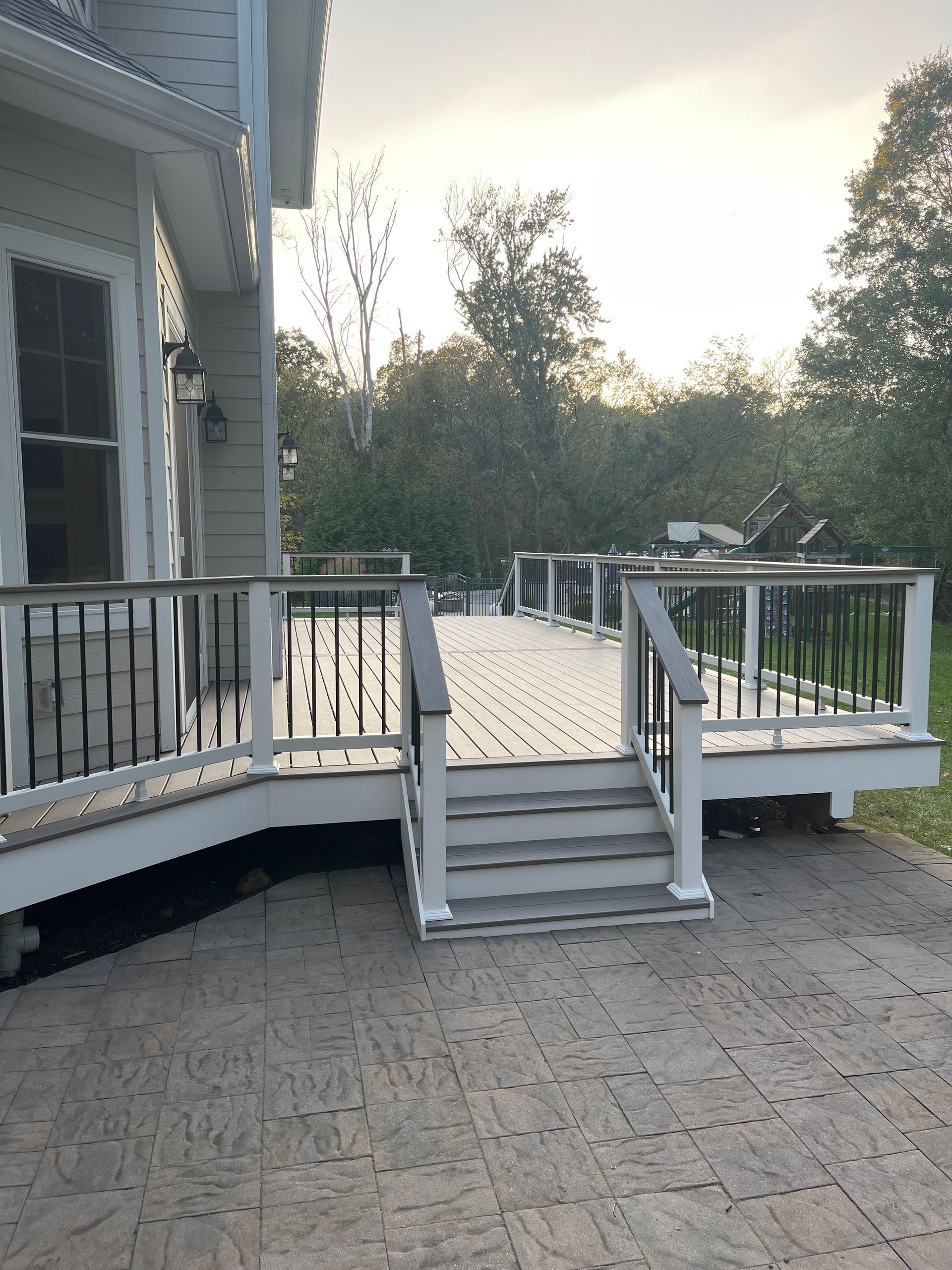 A wooden deck with stairs attached to a house. Gray decking, white railings, and a dark railing with a grassy area.