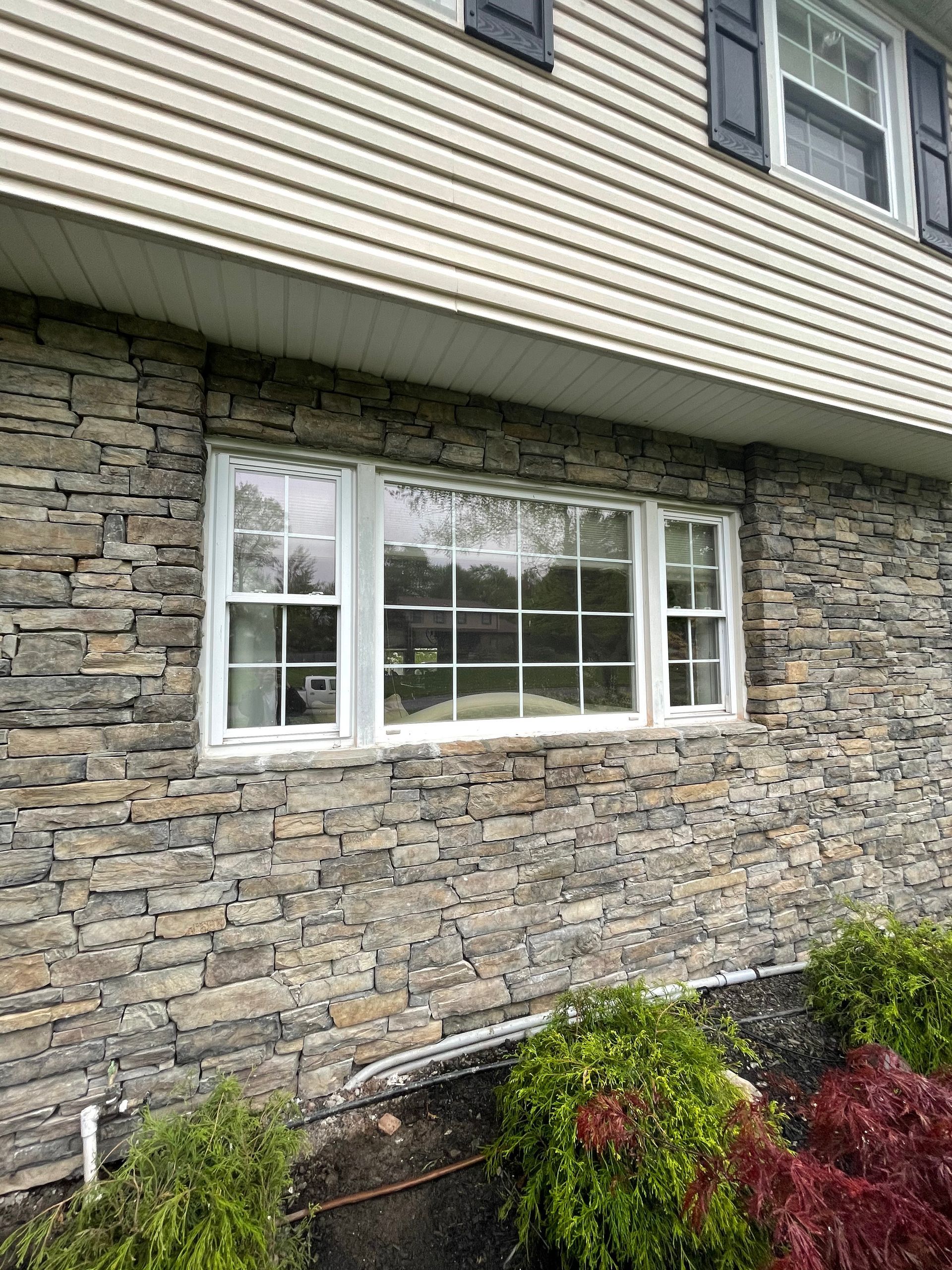 Stone-covered house exterior with a white-framed window. Green bushes in the foreground, and a light-colored siding on the upper part.