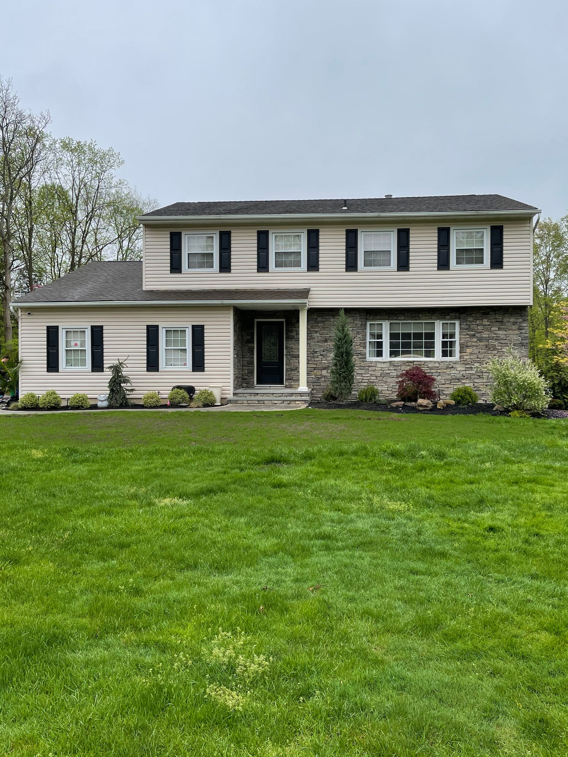 Two-story house with tan siding, black shutters, stone accents, and a large front lawn.