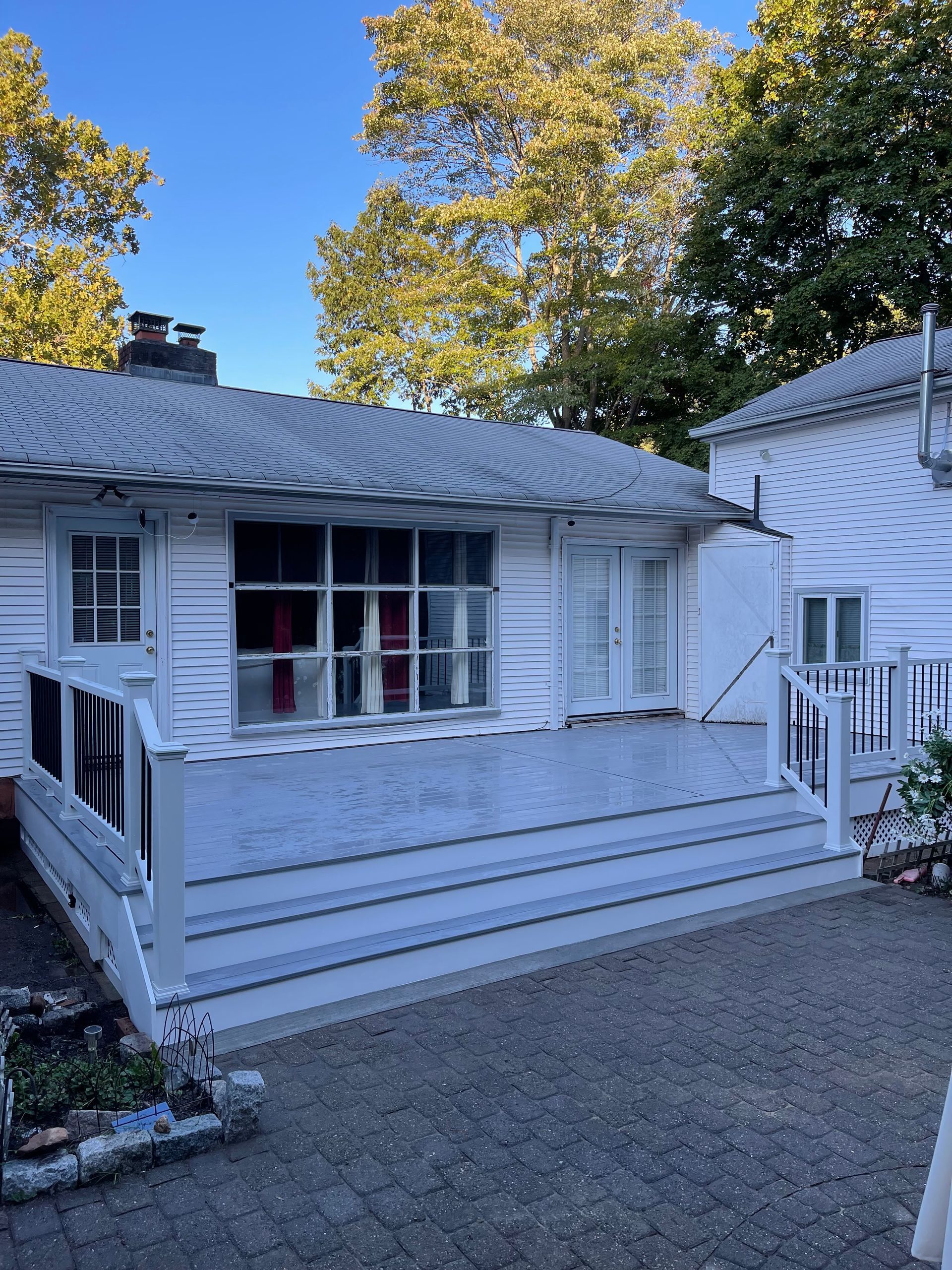 White house with deck and red curtains in window. Trees in the background, blue sky.