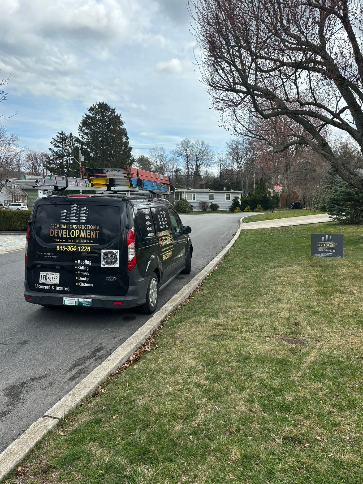 Black work van parked on a street with a sign on the lawn.