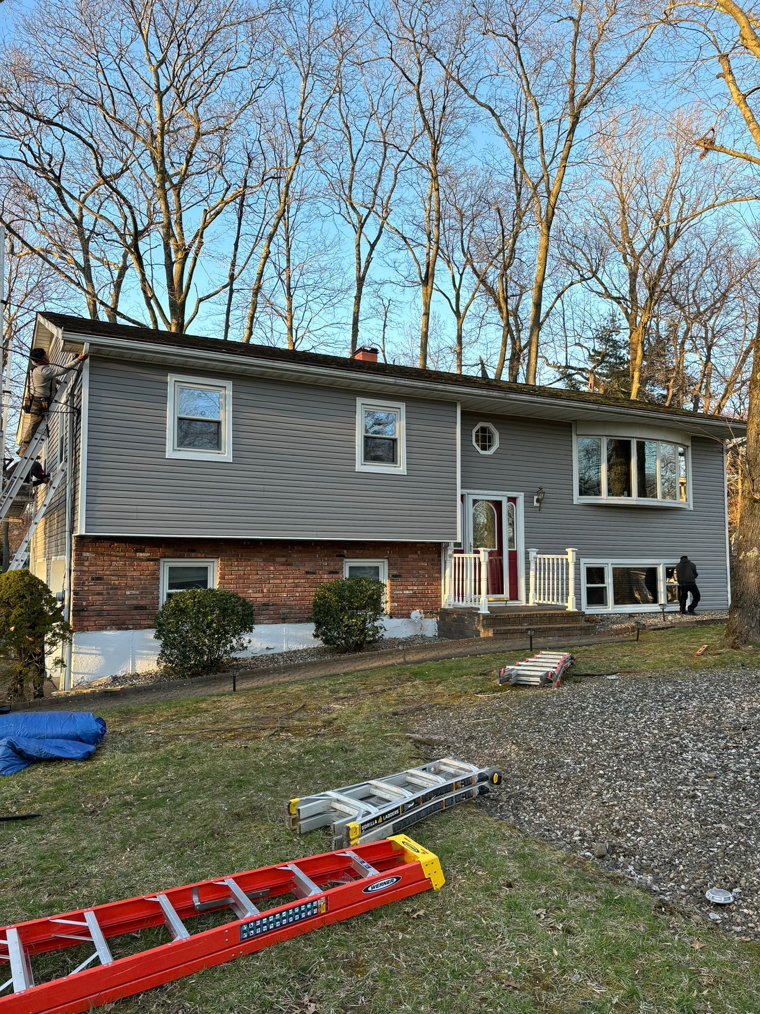 Two-story house with gray siding, brick base, and a red door. A ladder rests on the grass.
