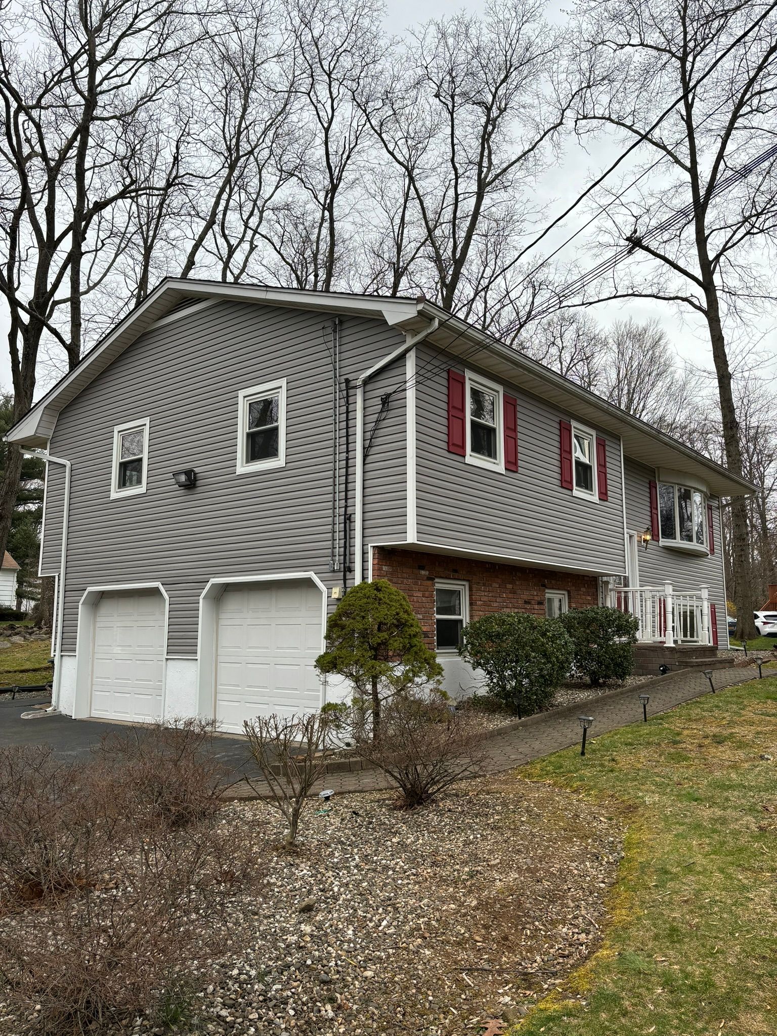 Gray two-story house with white garage doors, red shutters, and a sloped driveway lined with small plants.