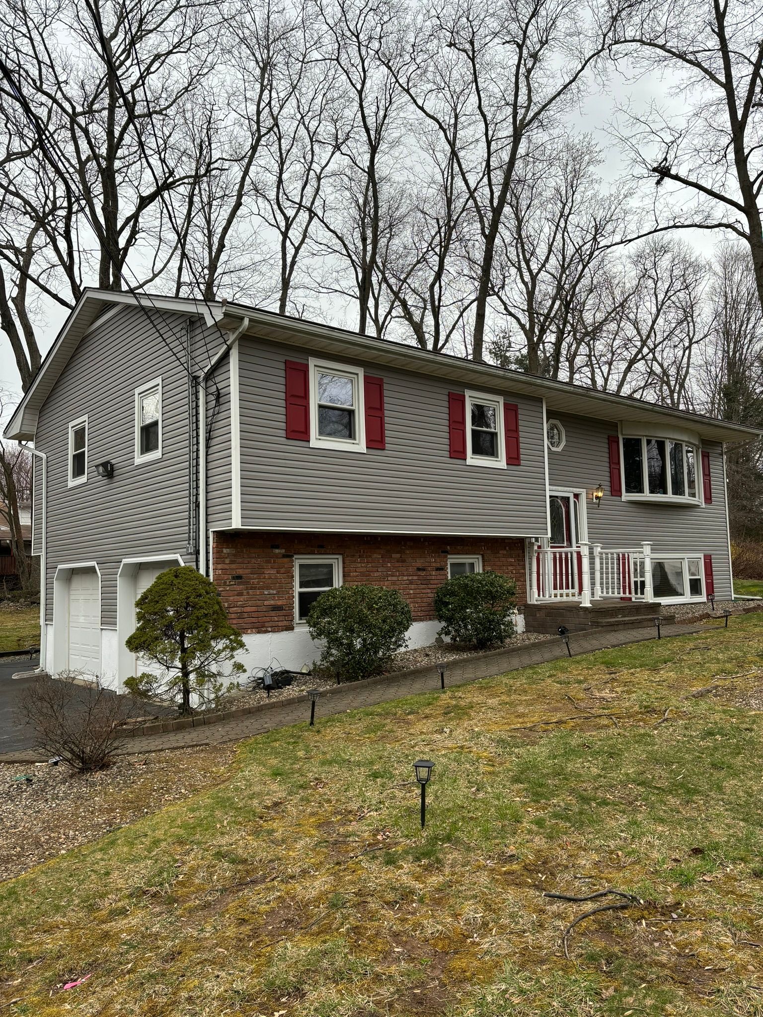 Two-story house with gray siding, red shutters, and a brick facade, set on a hill with sparse landscaping.