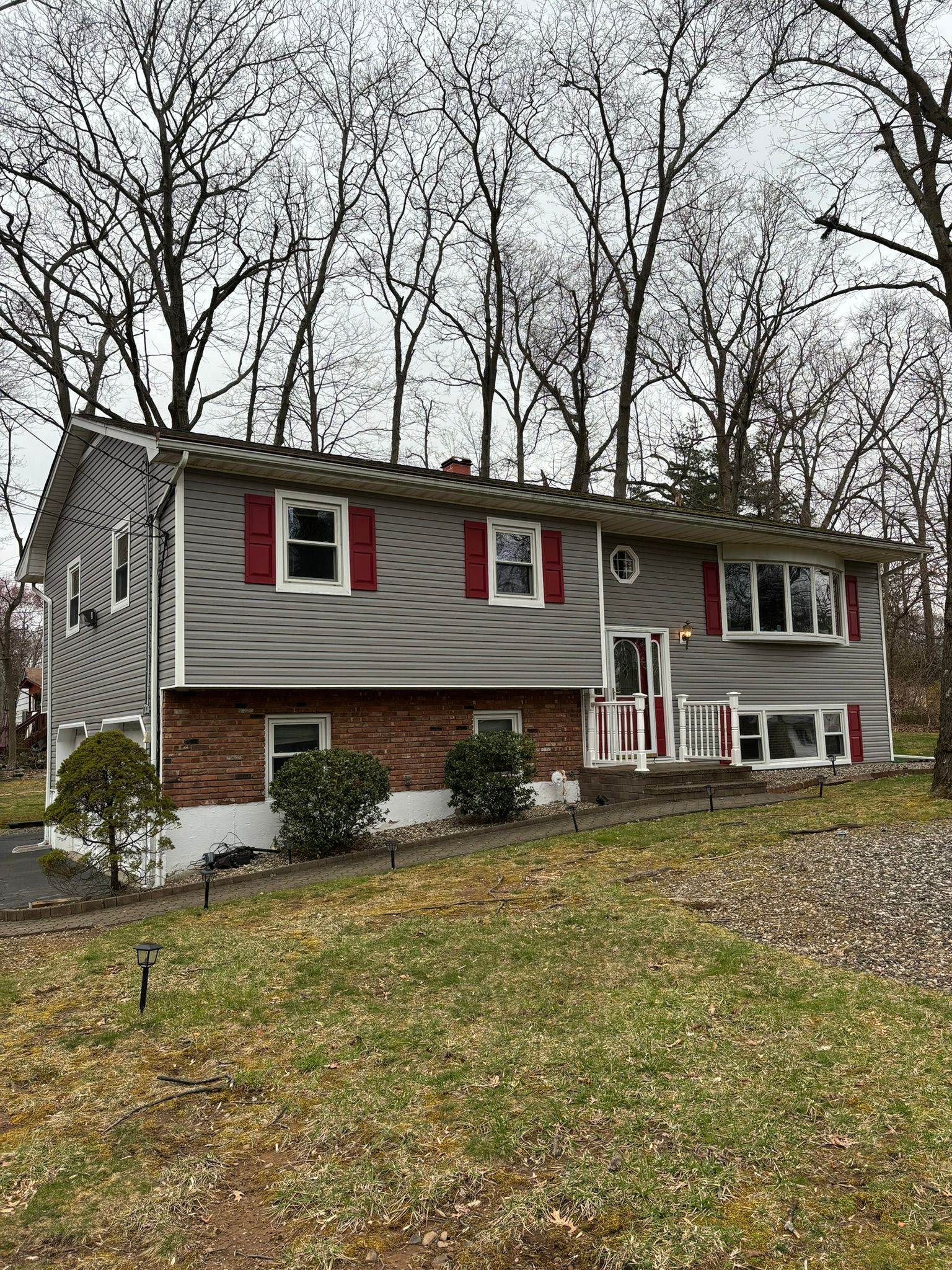 Two-story house with gray siding, red shutters, and a brick lower level. Trees in the background.