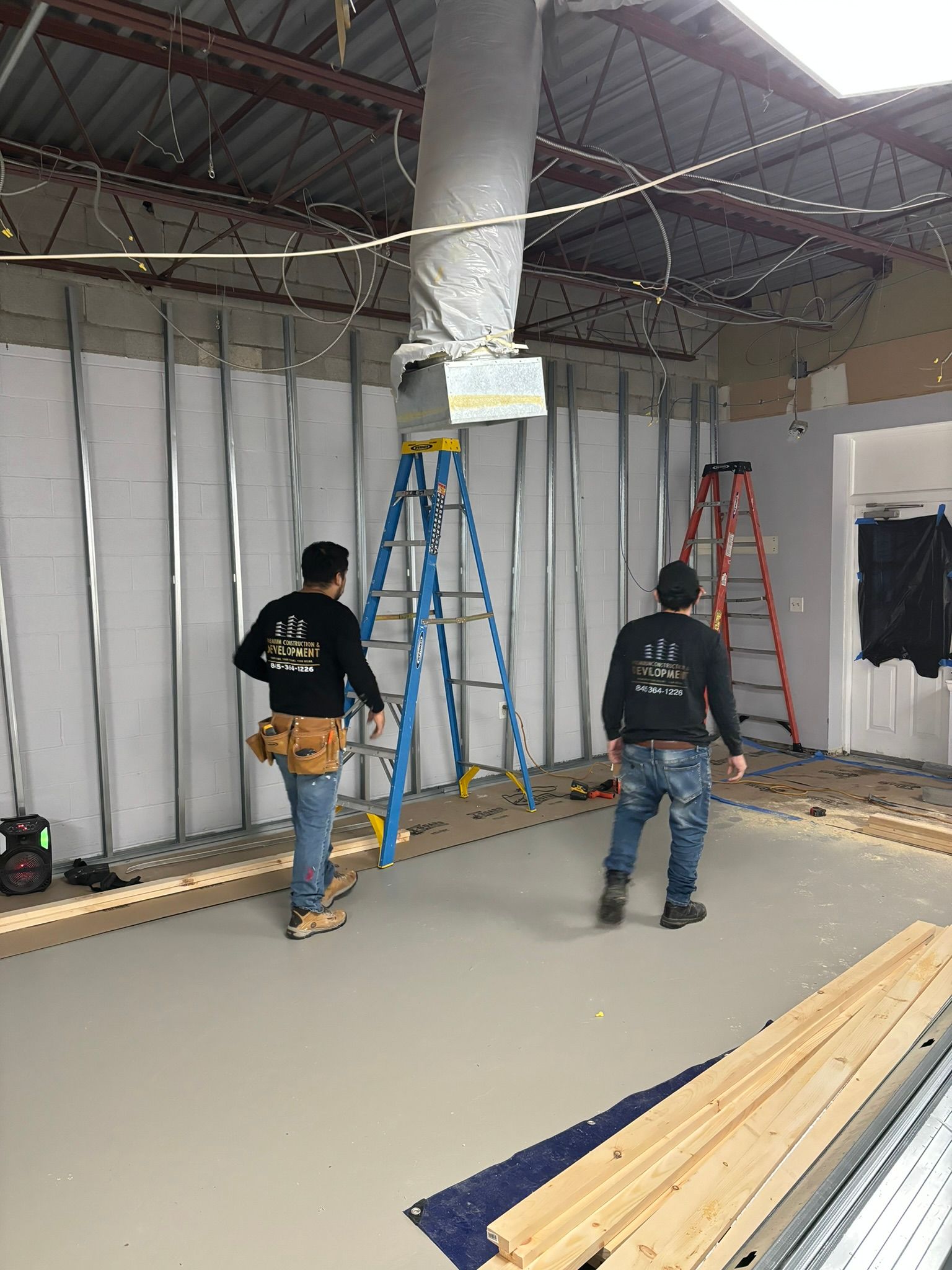 Two workers inside a construction site. One carries a ladder, the other looks on. Drywall and materials are visible.