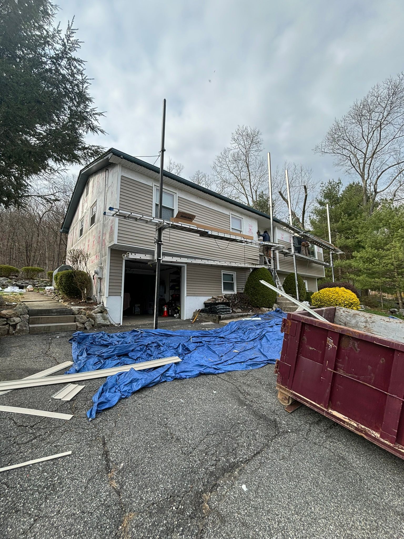 House exterior under construction, with scaffolding, tarp, and dumpster. Overcast sky.