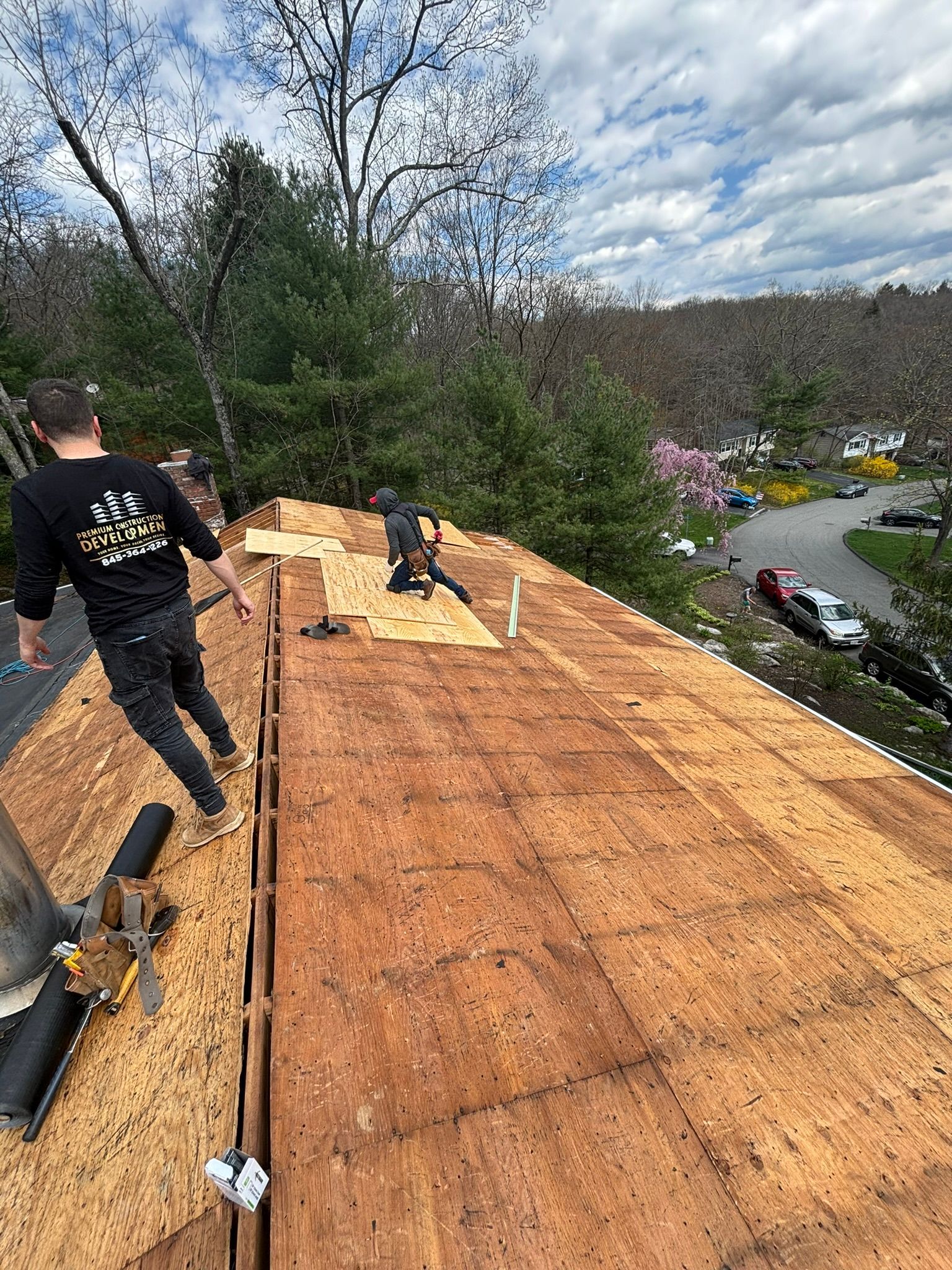 Two roofers on a rooftop, replacing wood panels. Overcast day with a view of trees and cars.