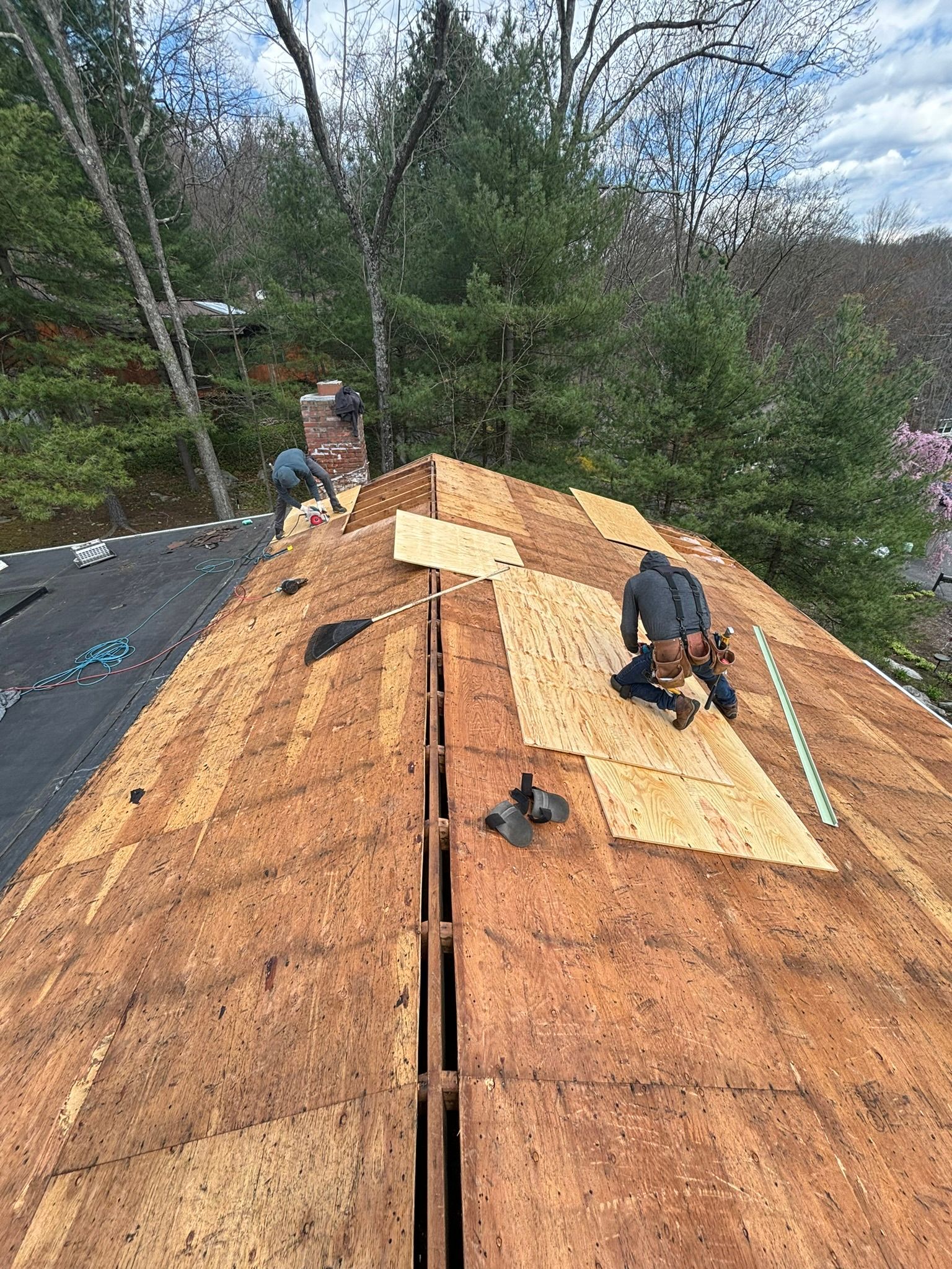 Roofers working on a house roof. One kneels, the other stands. Brown wood and tools. Outdoors.