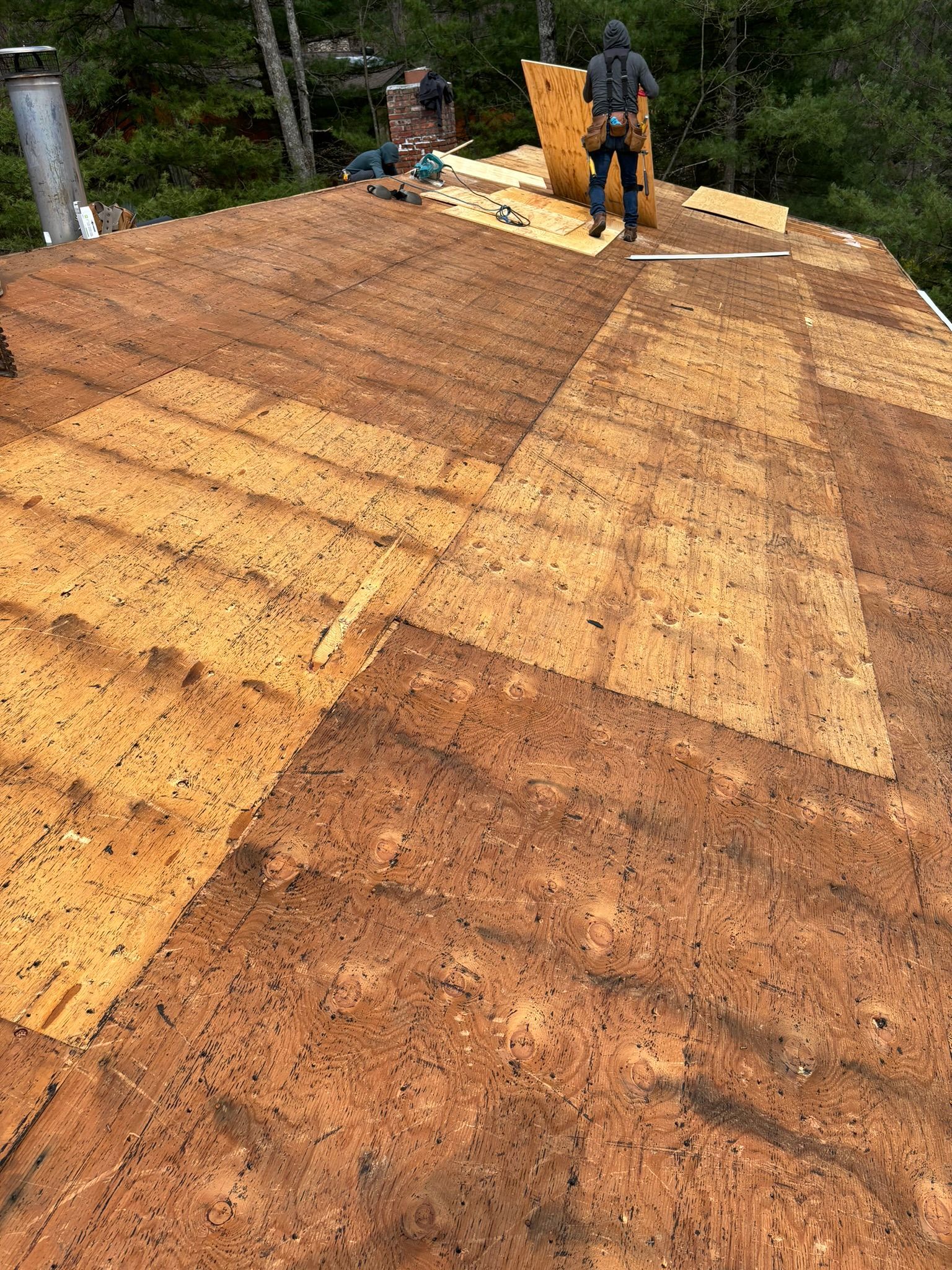Roofer on a sloped roof replacing wooden sheathing, surrounded by tools and materials.