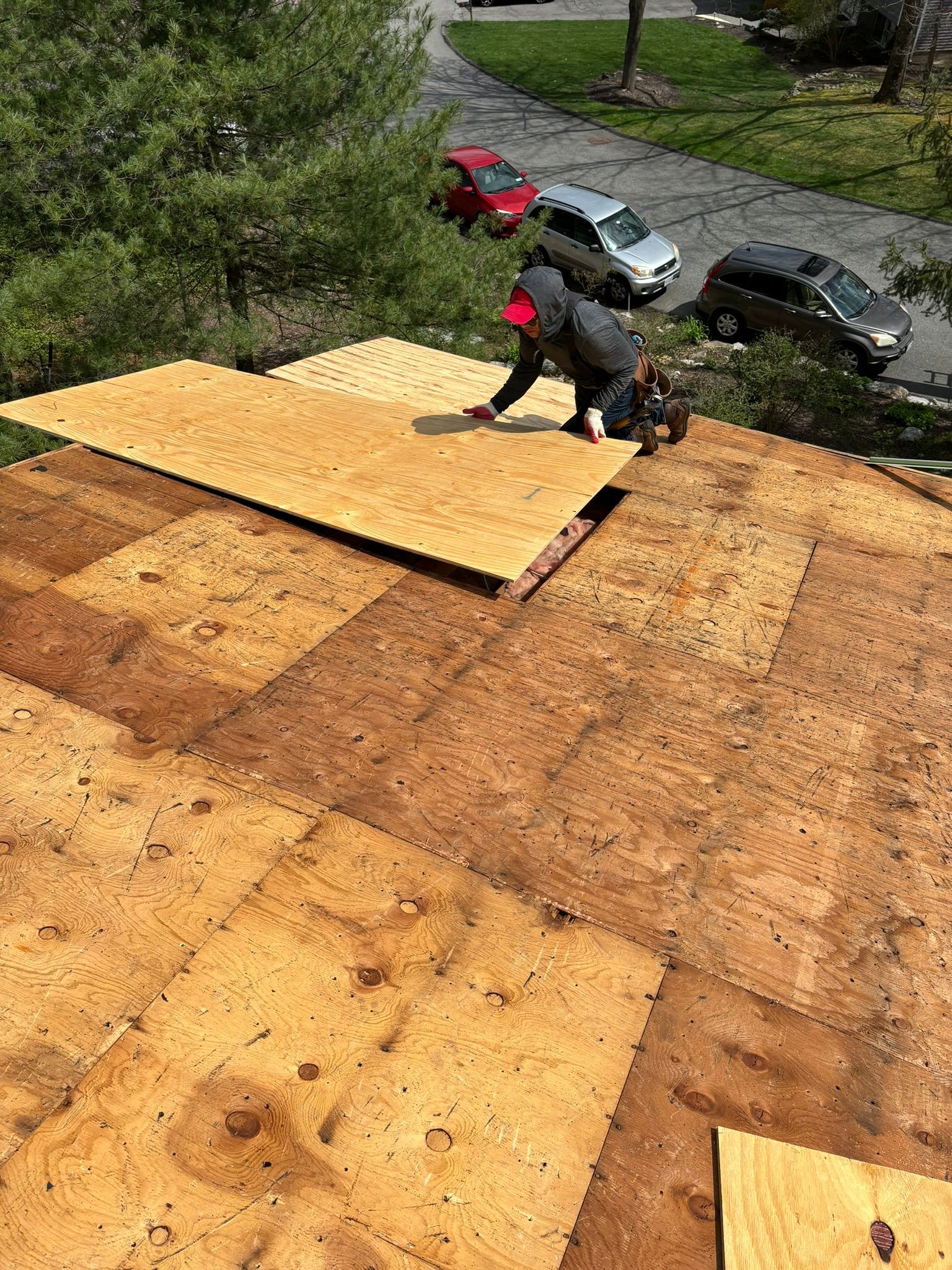 A person installing plywood on a roof; cars and trees in the background.