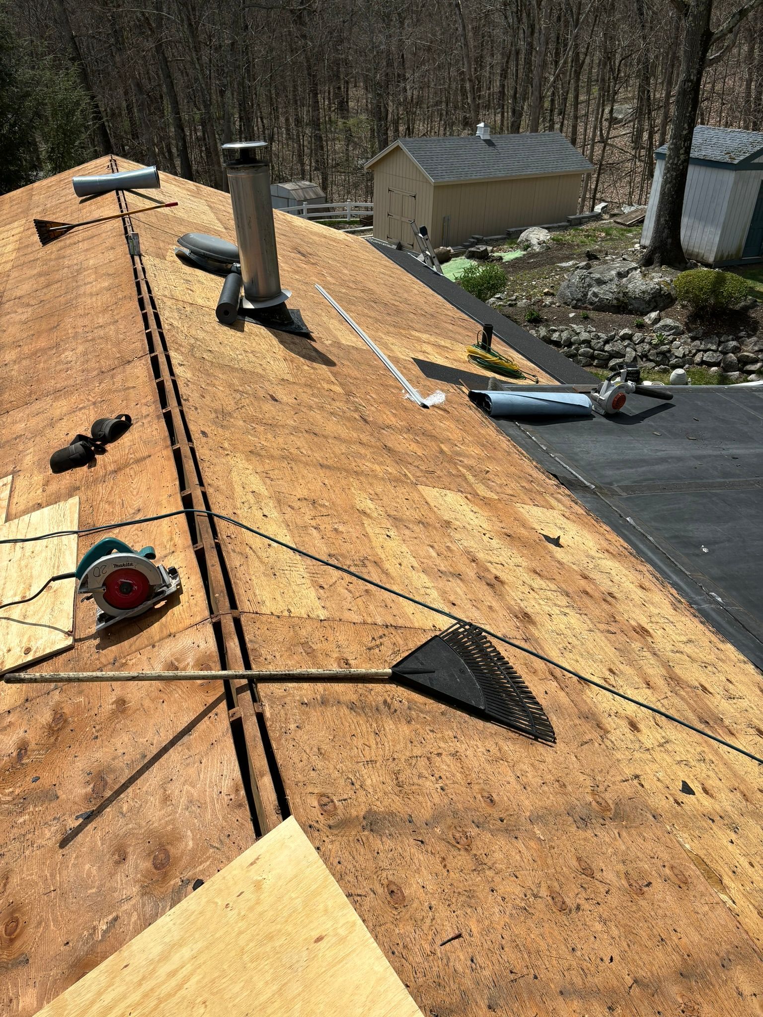 Roof with exposed plywood, tools, and chimney during construction, with buildings and trees in the background.