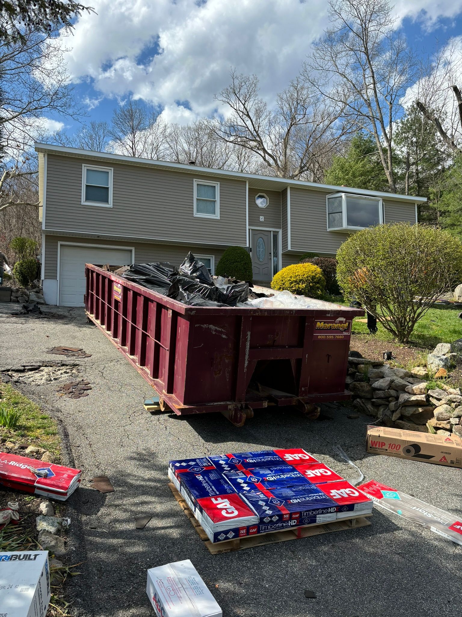 A home with a large dumpster in the driveway, construction supplies nearby, and a partially cloudy sky.