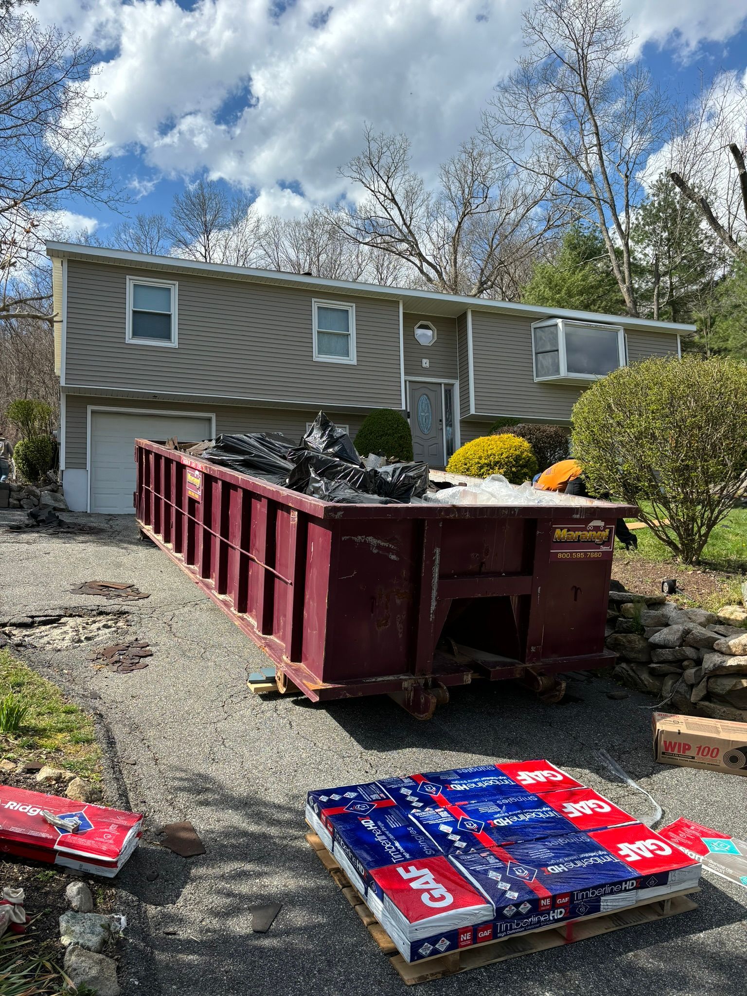 House with a large dumpster in the driveway and stacked roofing materials. Sunny day, residential setting.
