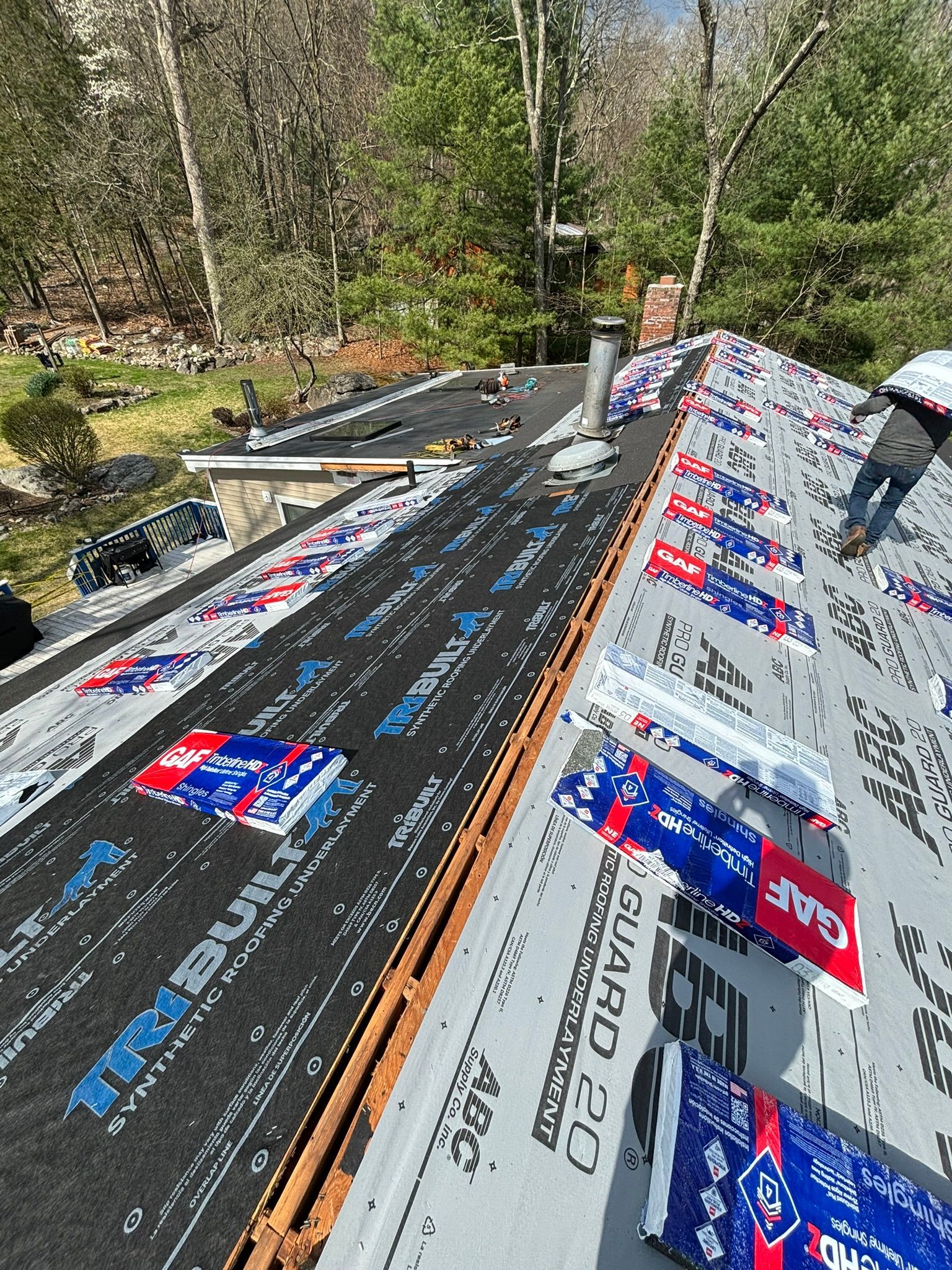Roofer working on a house roof. Underlayment and shingles are being installed on a sunny day in a wooded area.