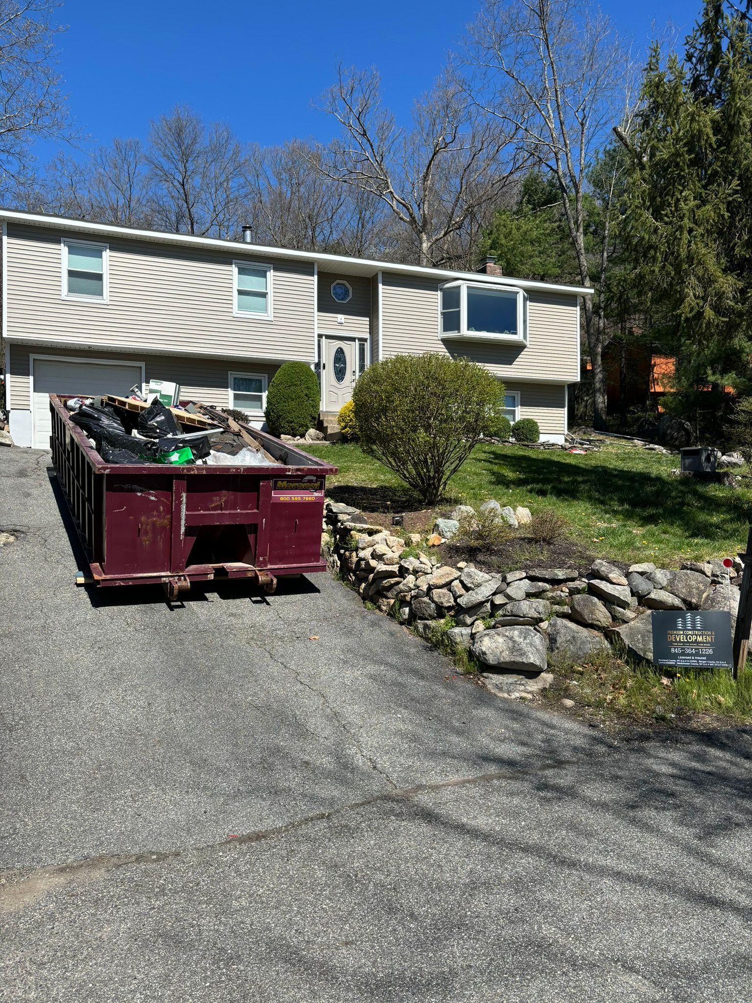 A burgundy dumpster in a driveway in front of a light-colored house. Green grass and trees are in the background.
