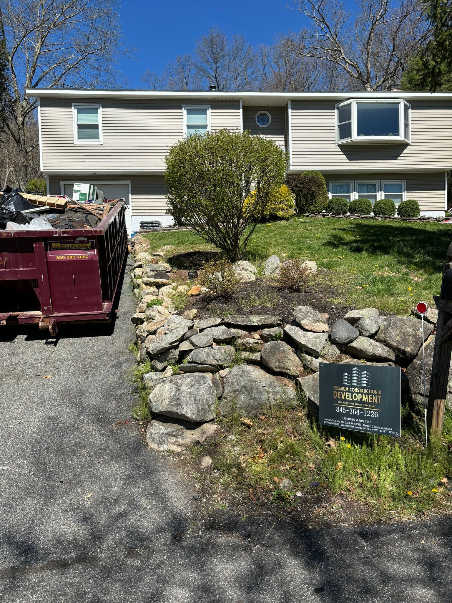 House with a rock wall in front, dumpster in driveway, sunny day.