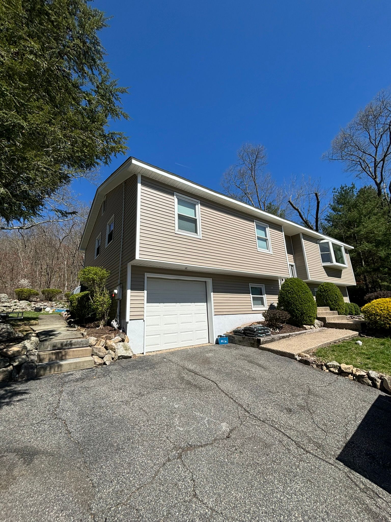 Two-story beige house with a driveway. Garage door, windows, and trees under a clear blue sky.