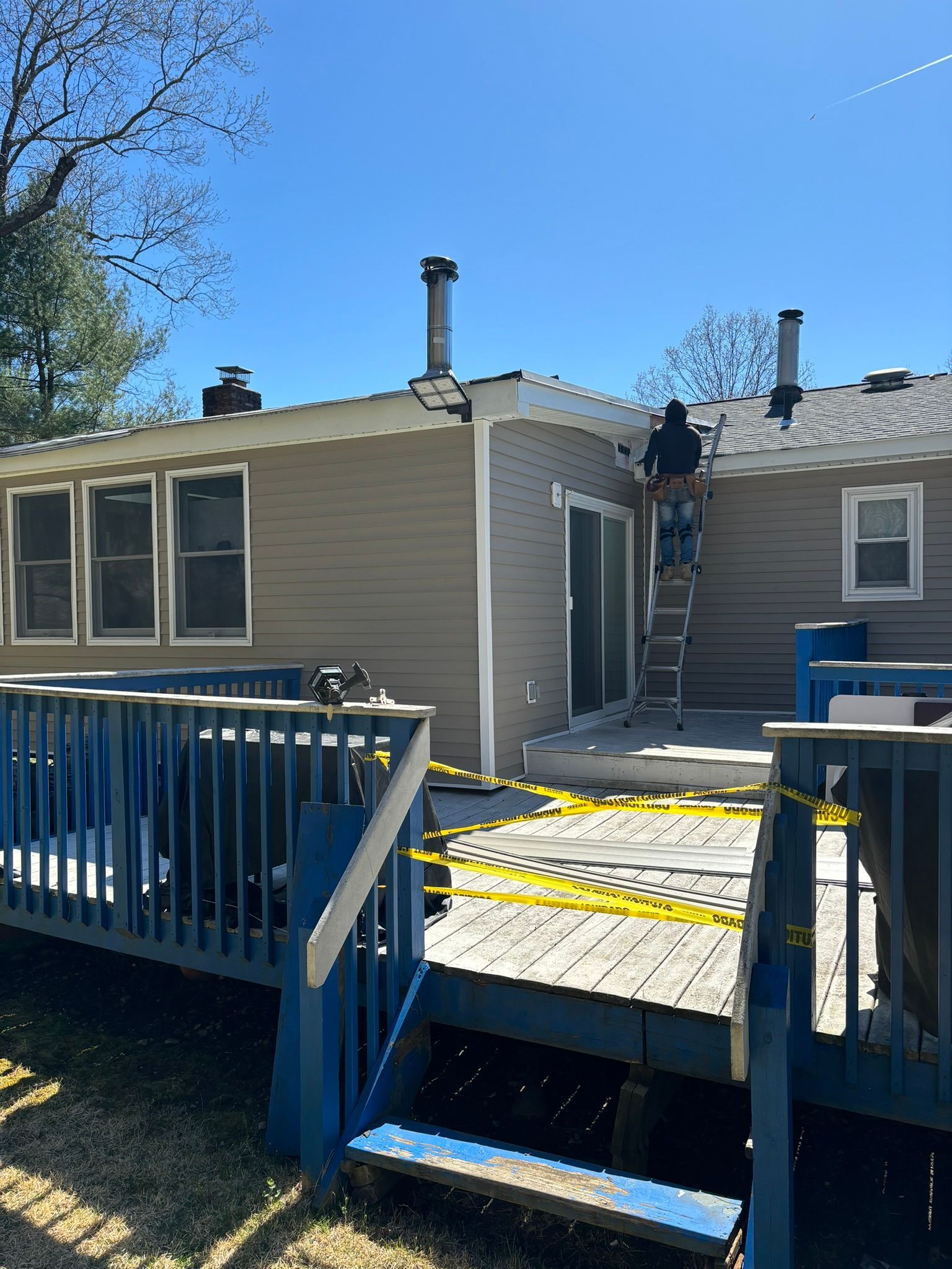 Person on a ladder working on a house roof. Blue deck in foreground, yellow caution tape, and clear sky.