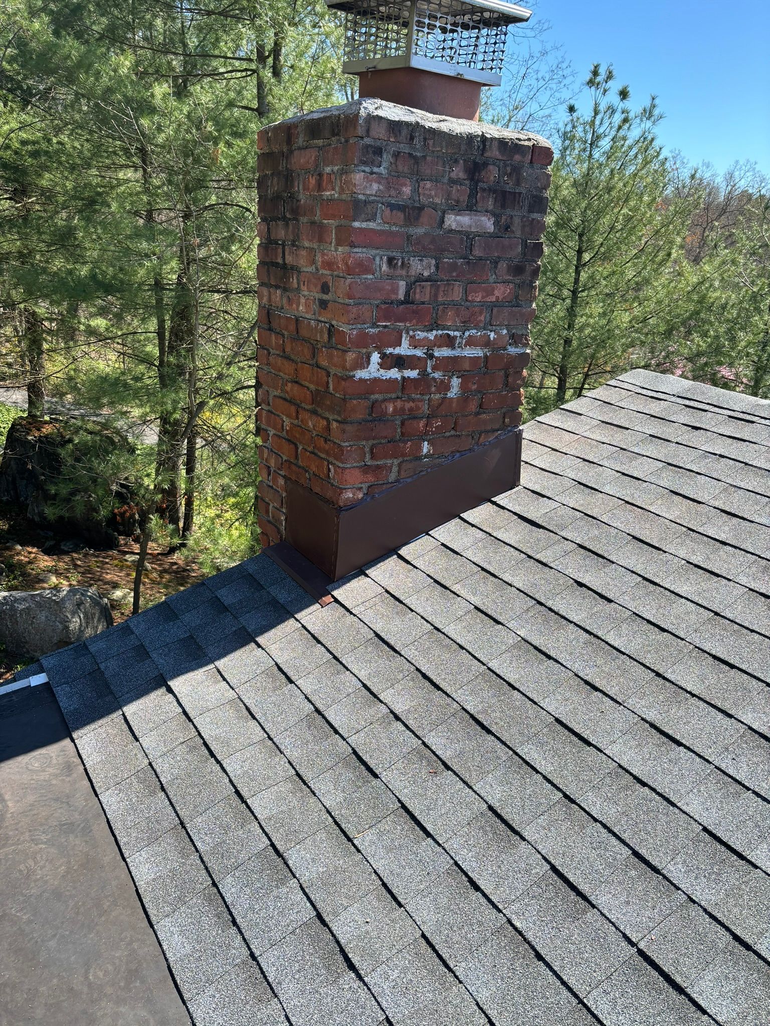 Brick chimney on a shingled roof with visible damage, metal flashing, and a capped top.