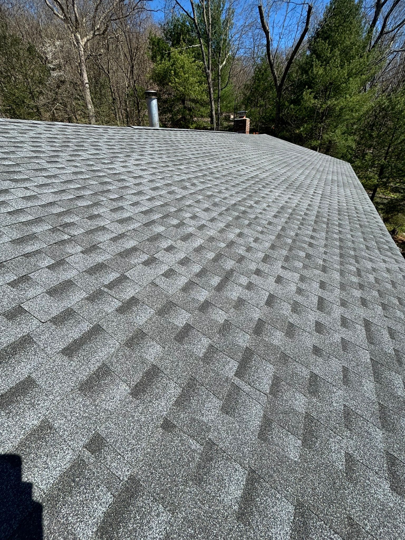 Gray asphalt shingle roof on a house, with a chimney and trees in the background.