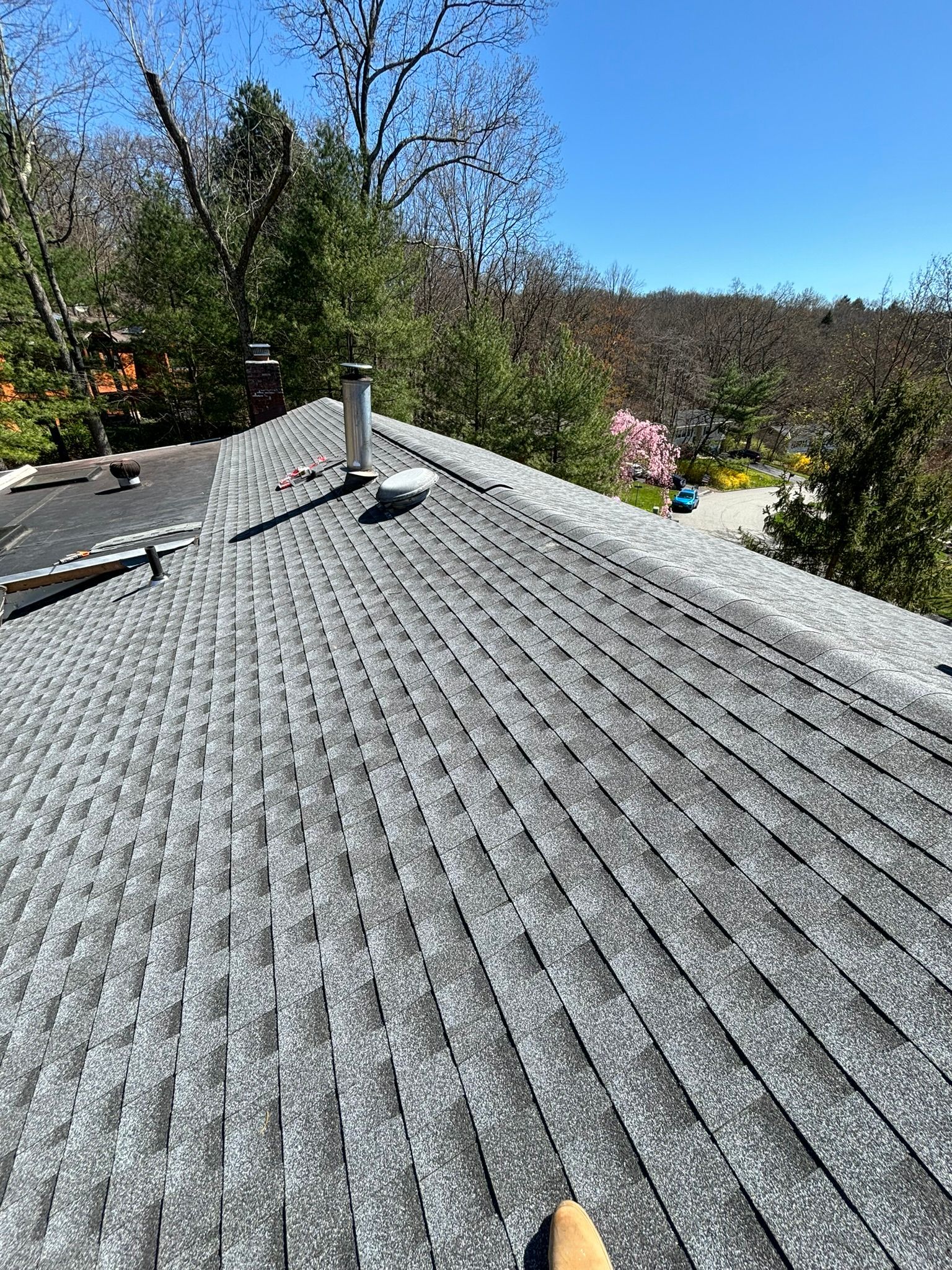Gray asphalt shingle roof on a house, with a metal vent pipe and trees in the background on a sunny day.