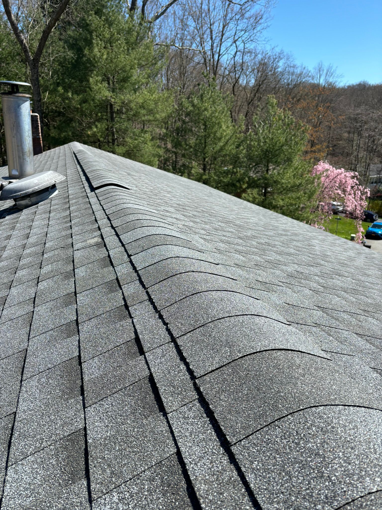 A shingled roof with a chimney and trees in the background under a blue sky.