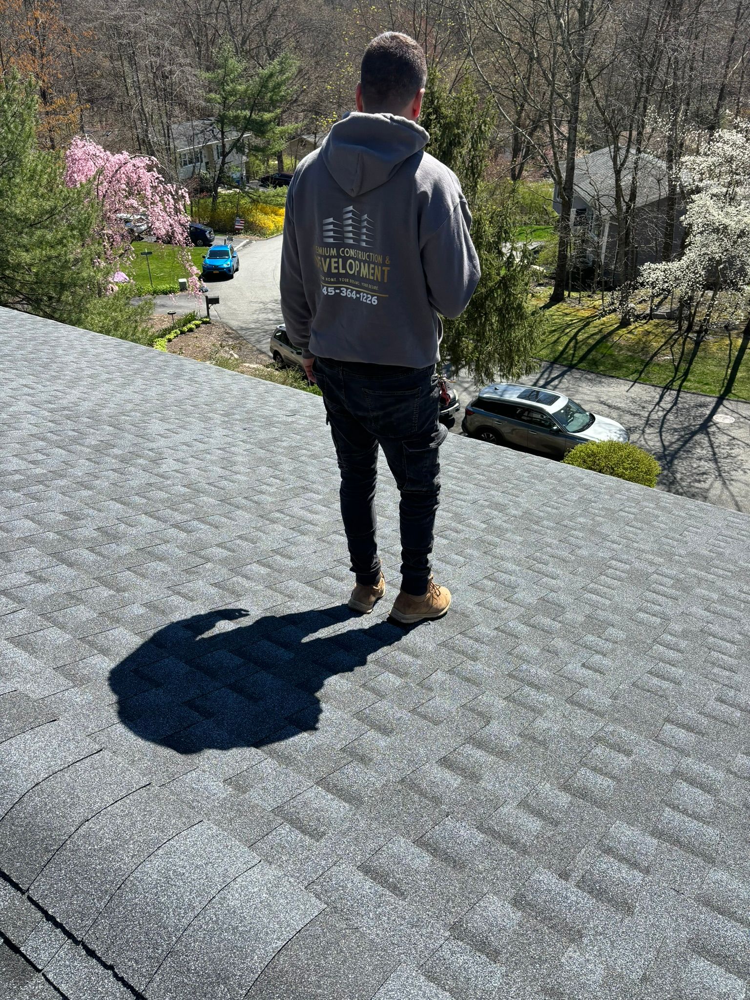 Man standing on a gray shingled roof, casting a shadow, with houses and trees in the background.